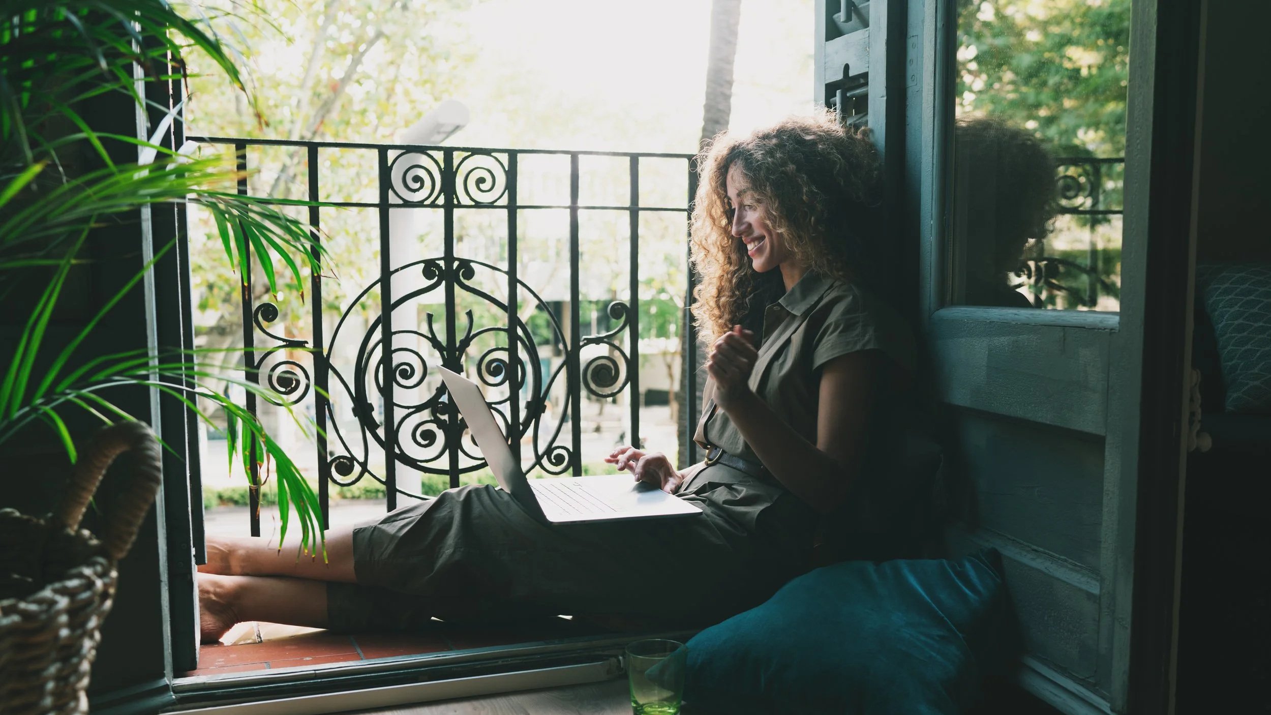 Woman with curly hair sitting with legs extended on a balcony, smiling while looking at a laptop, surrounded by green plants and trees.