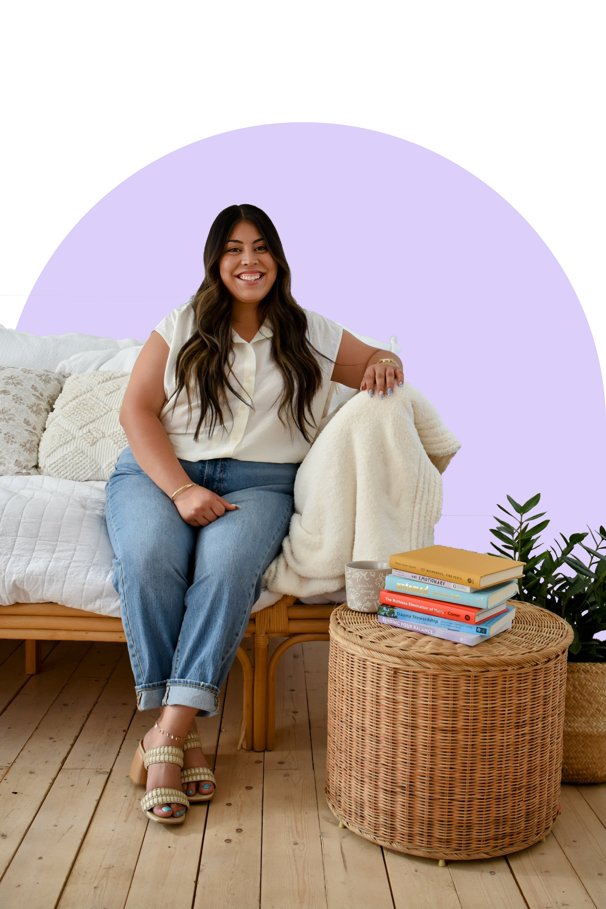 A woman with long dark hair and a white shirt sitting on a couch, smiling. She is wearing blue jeans and cream-colored platform sandals. Next to her is a woven round side table with a stack of colorful books and a plant in the background.