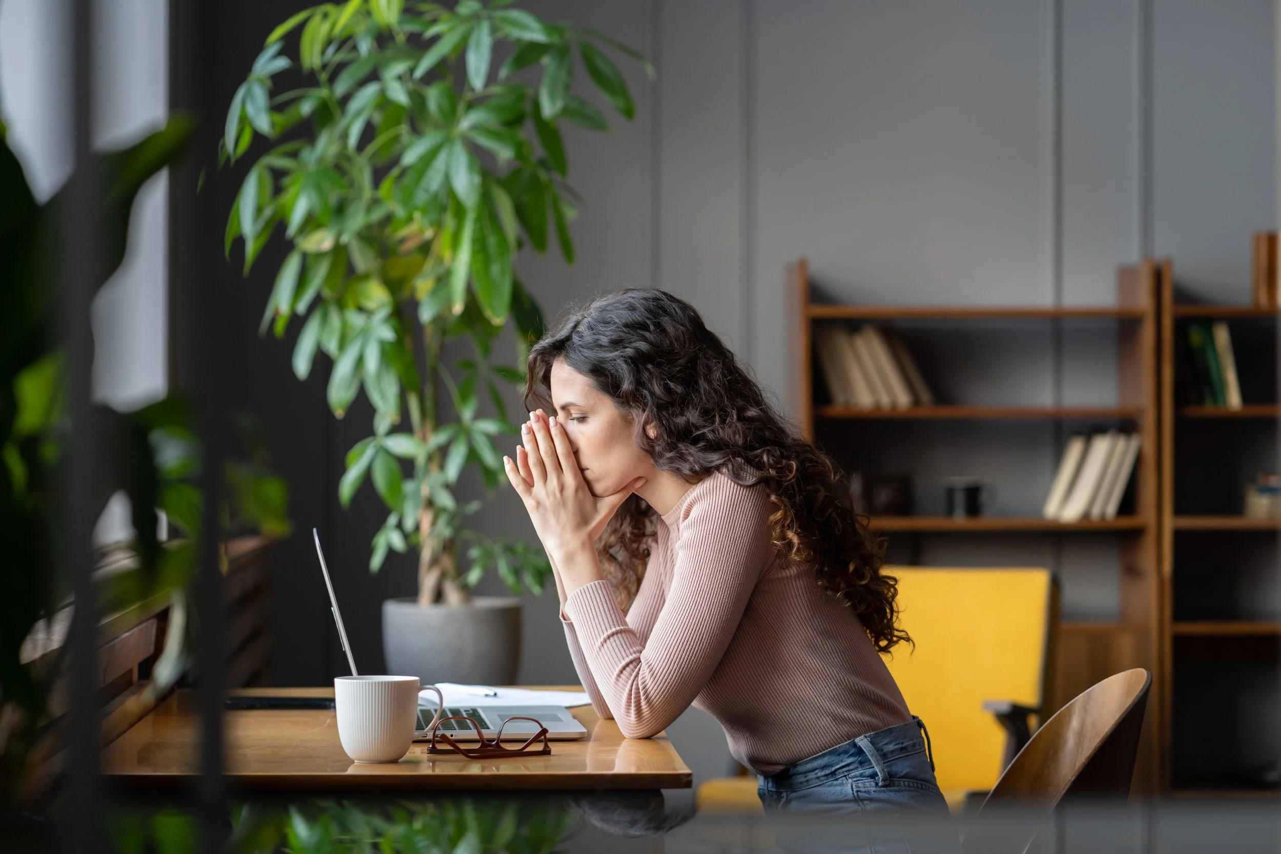 A woman with curly hair and light skin, sitting at a wooden table with a laptop, notebook, and coffee mug, appears stressed with her hands covering her face in a modern office or library setting with bookshelves and green plants.