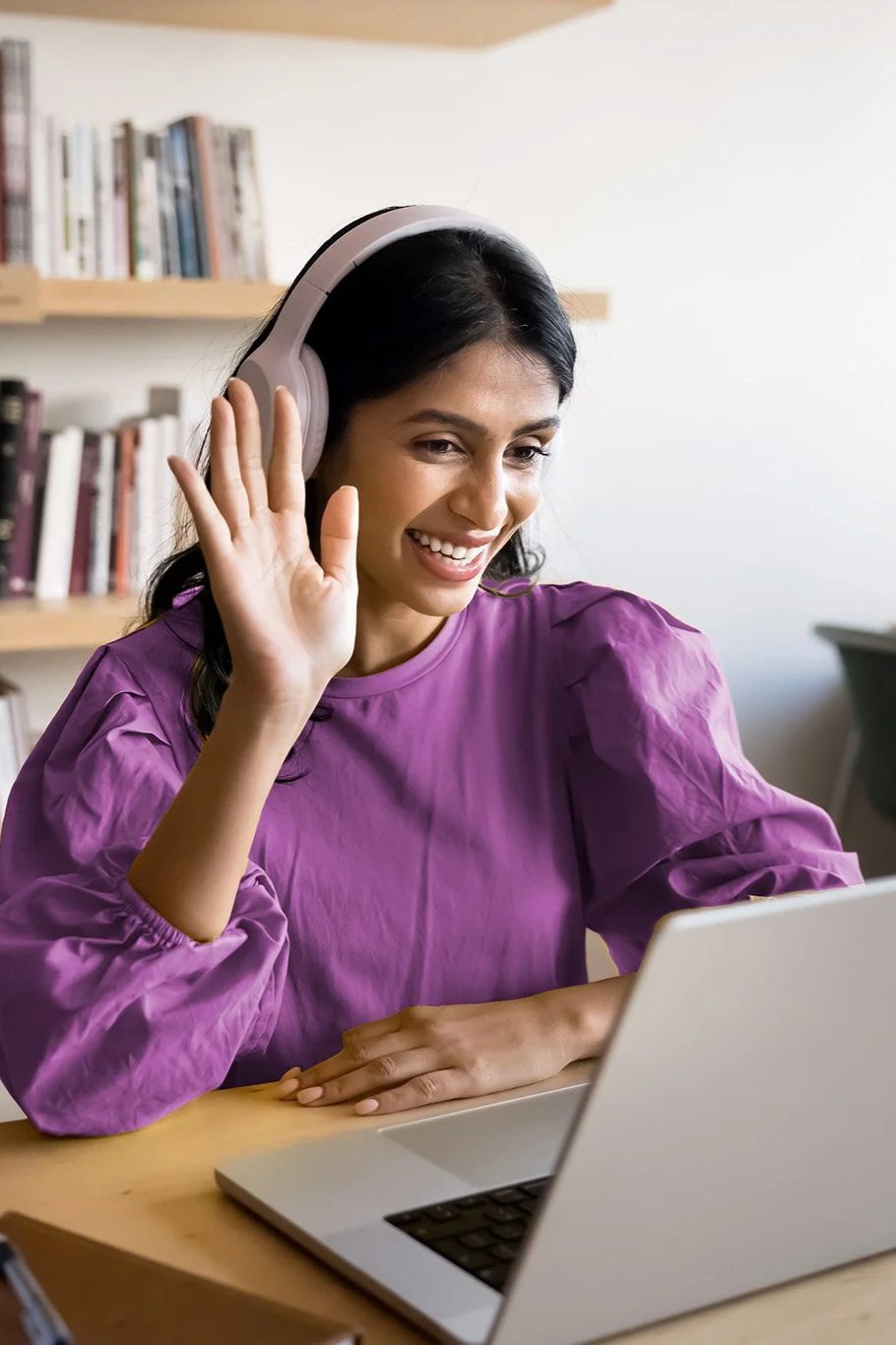 A woman with dark hair in a purple blouse wearing over-ear headphones, smiling, waving, and looking at a laptop screen in a room with a wooden bookshelf in the background.