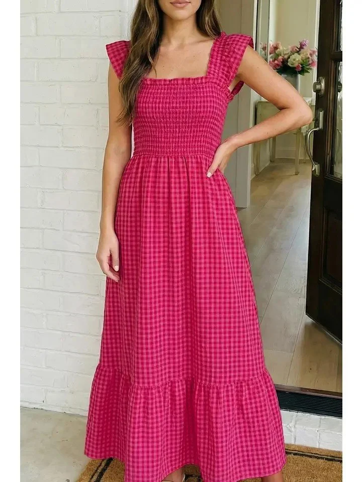 Woman wearing a pink, checkered, sleeveless maxi dress with ruffled shoulder details, standing indoors near a white brick wall and a door with a flower arrangement visible in the background.
