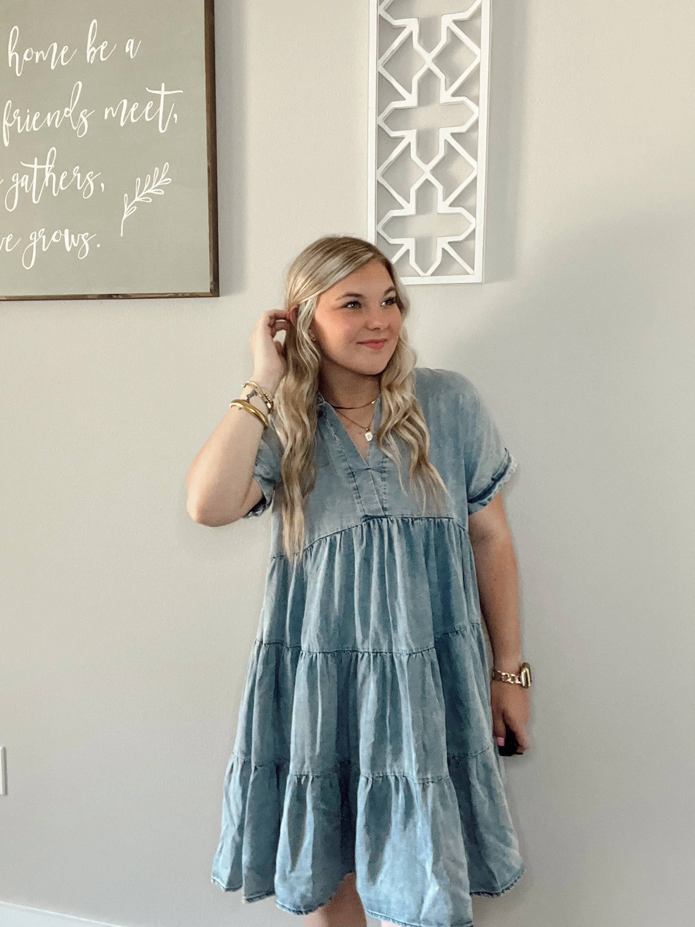 A young woman with long, wavy blonde hair wearing a loose denim dress, standing in front of a beige wall decorated with framed wall art. She is smiling with her head slightly tilted, touching her hair with her right hand, and wearing jewelry including bracelets, a watch, and layered necklaces.