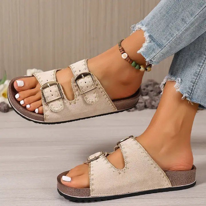 Person wearing beige slip-on sandals with two adjustable buckles, frayed hem jeans, and a beaded bracelet on ankle.