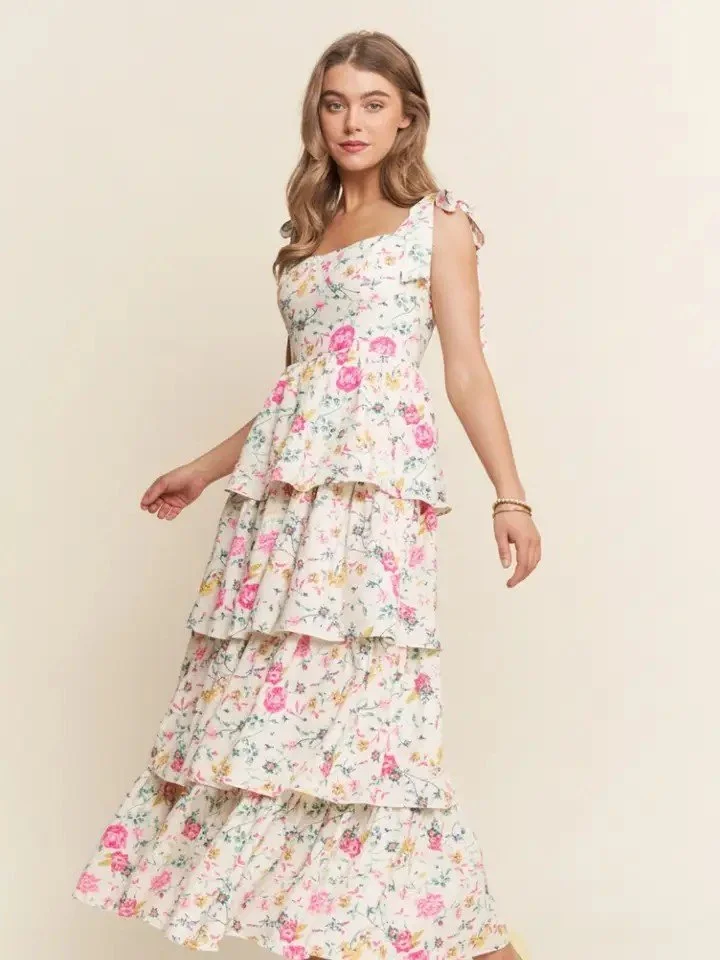 A woman in a sleeveless white floral maxi dress with pink and yellow flowers and tiered ruffles, standing against a plain beige background.