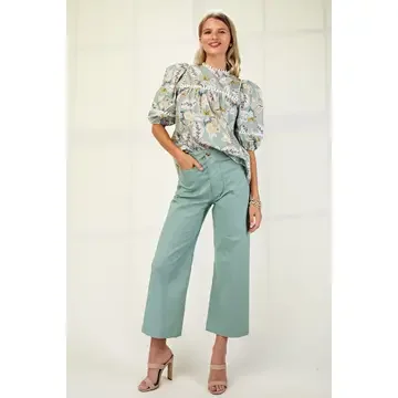 Woman standing and smiling, wearing a floral blouse, mint green wide-leg trousers, and beige high heels.
