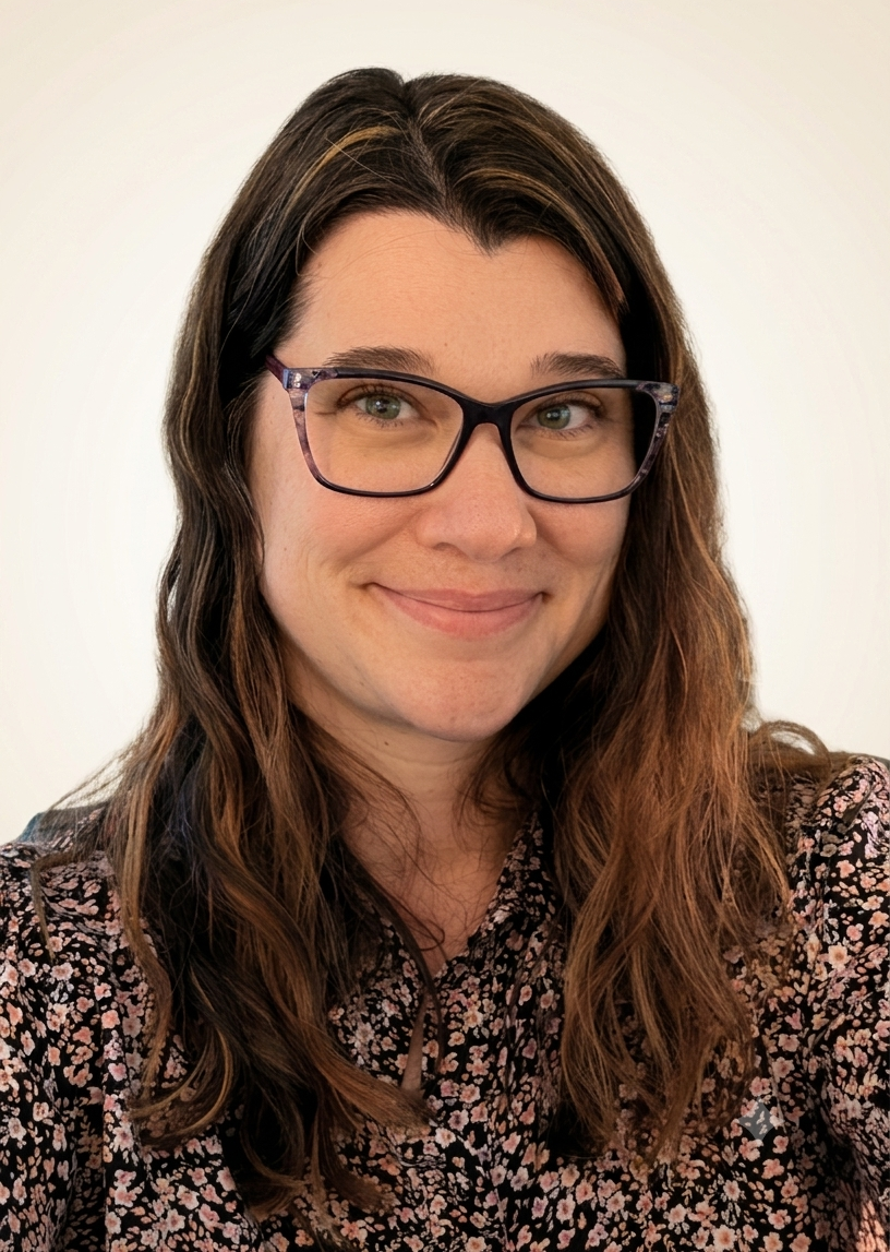 A woman with long wavy brown hair, glasses with purple frames, and wearing a floral blouse, smiling at the camera against a plain white background.