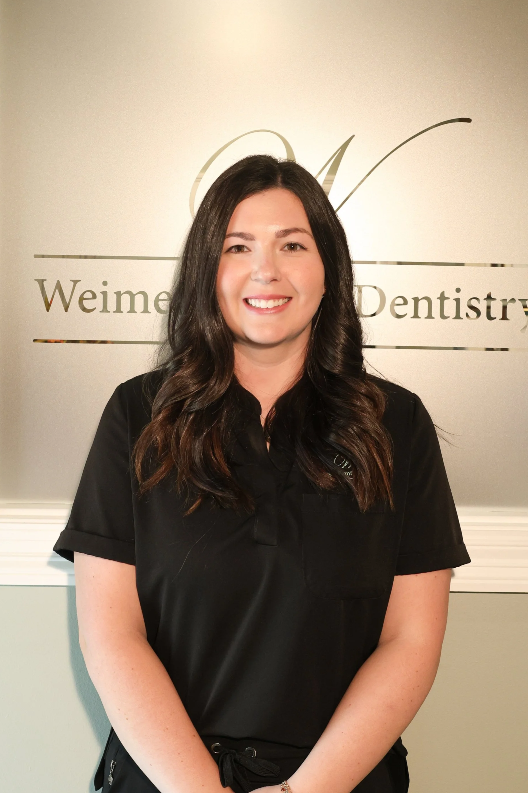A woman with long brown hair smiling in front of a glass wall with a logo and text, wearing a black scrub top.