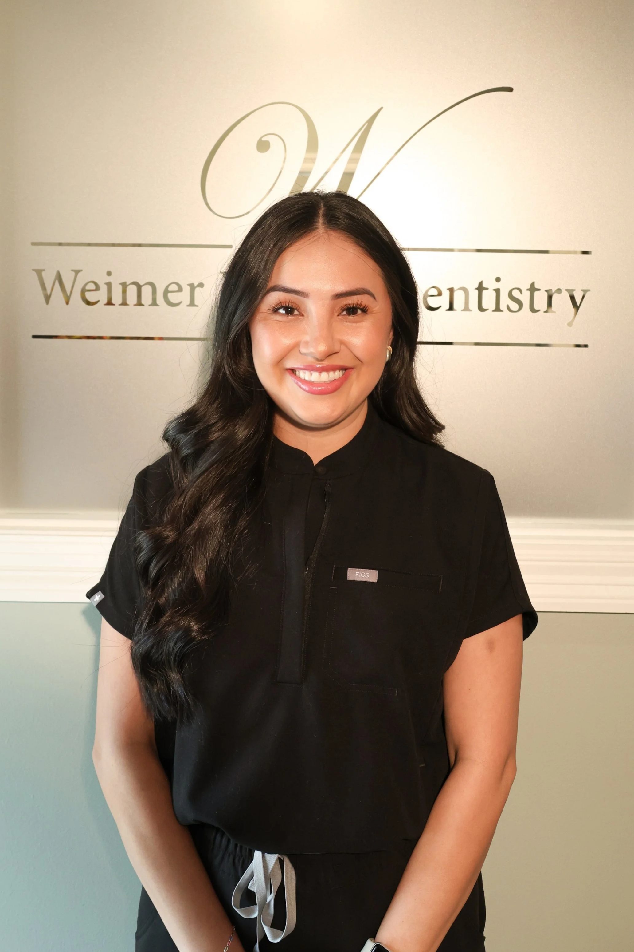 A woman smiling in a black medical uniform standing in front of a sign that reads 'Weimer Family Dentistry'.