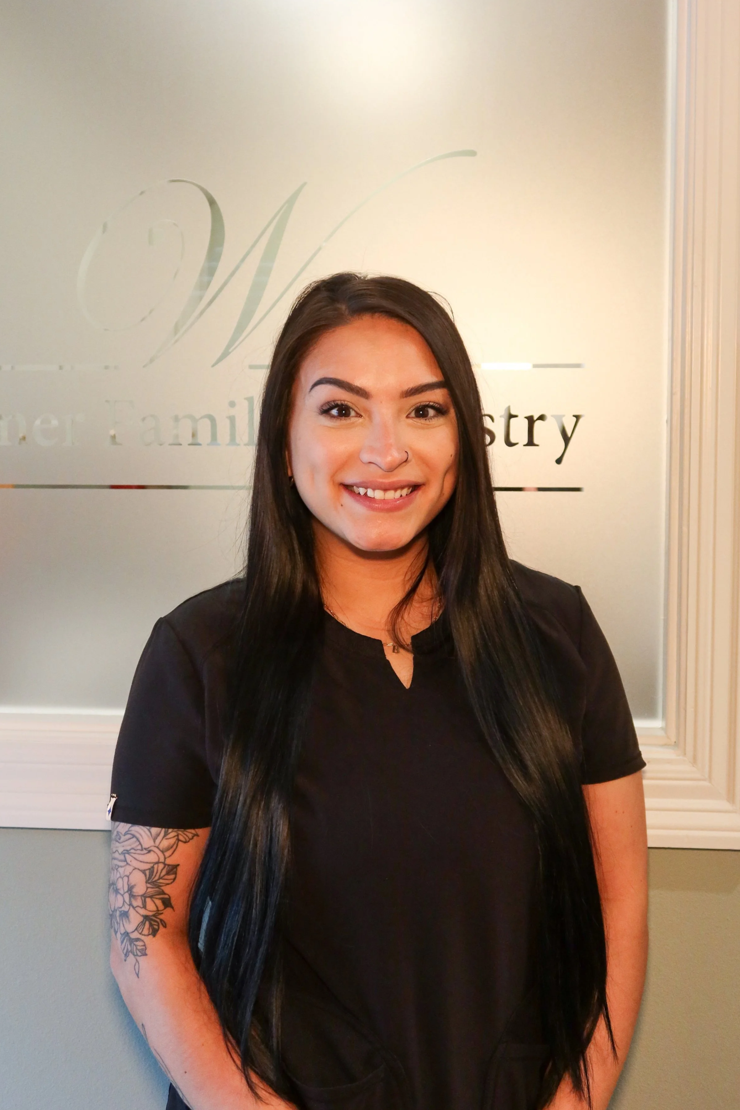 Smiling woman with long black hair, nose ring, and floral tattoo on her left arm, wearing a black top, standing indoors in front of frosted glass with a logo.