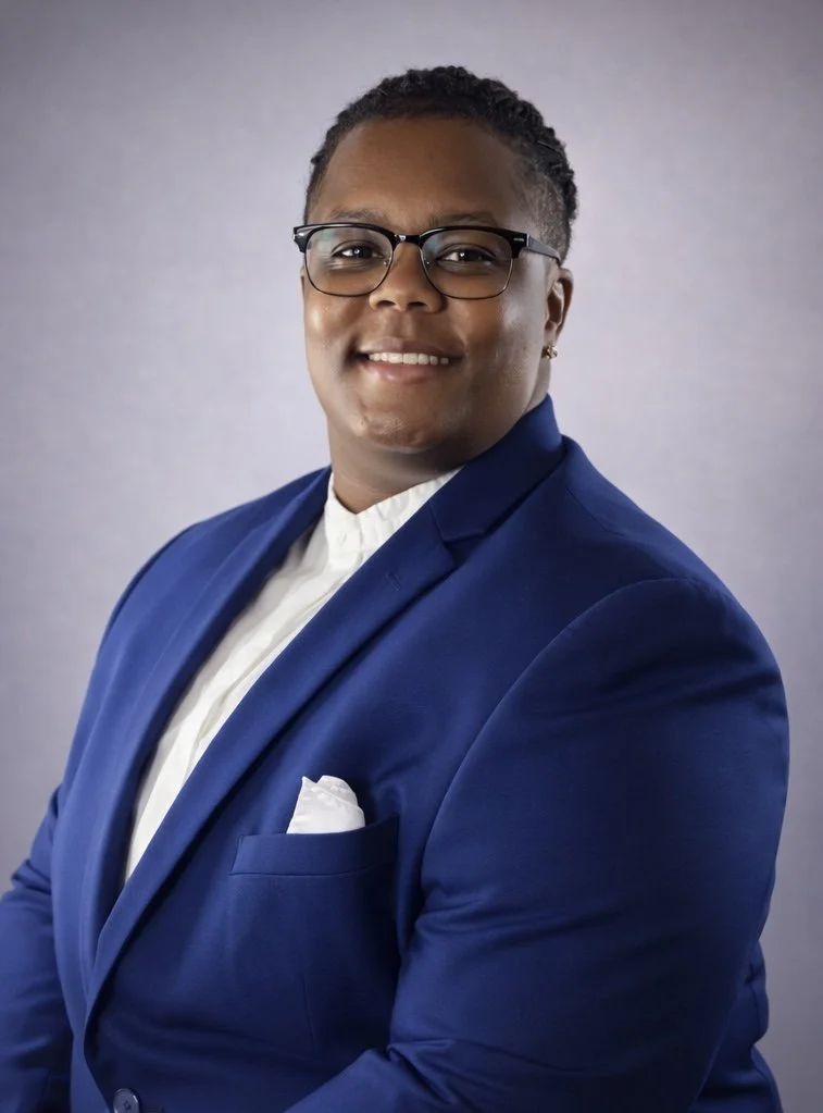 A person in a blue suit with a white shirt and pocket square, wearing glasses, smiling against a gray background.