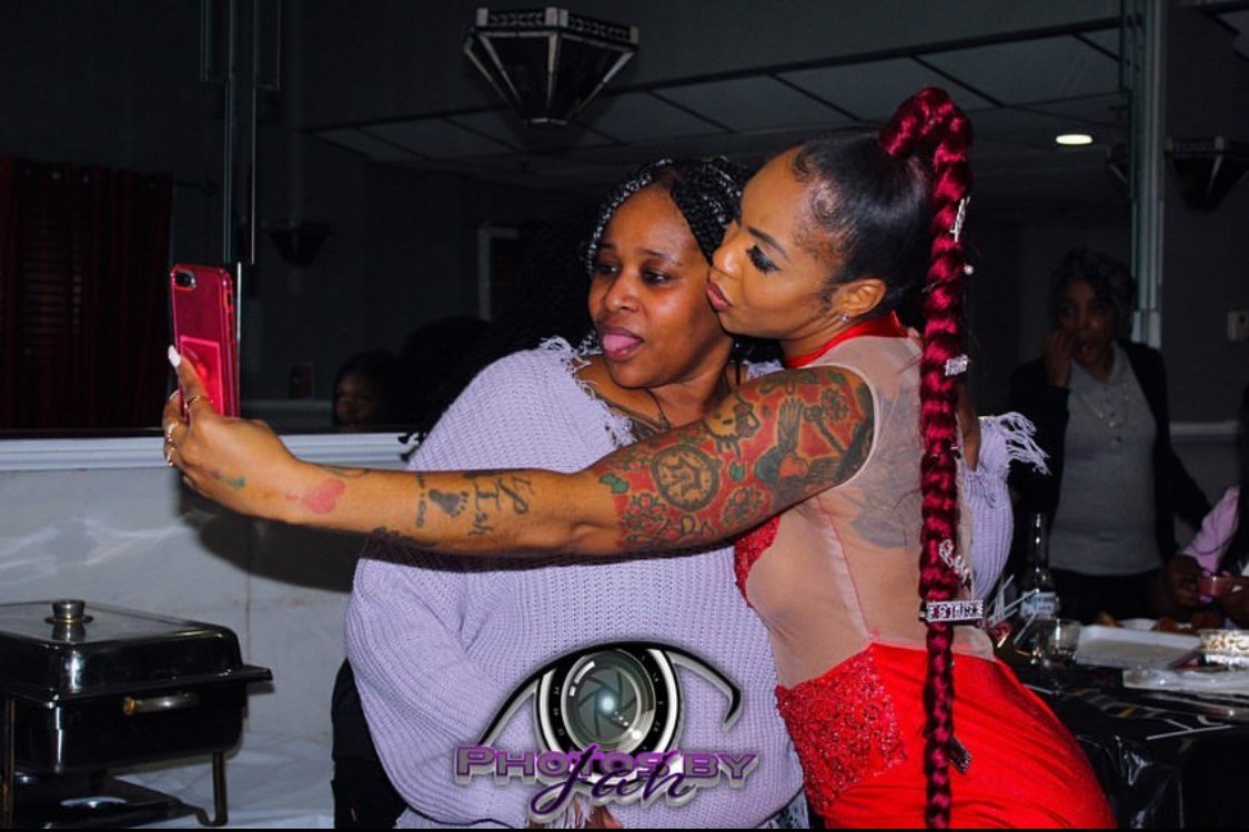 Two women taking a selfie together at an indoor gathering, one with a long red braid and tattoos, the other in a light purple sweater, both smiling and close to each other.