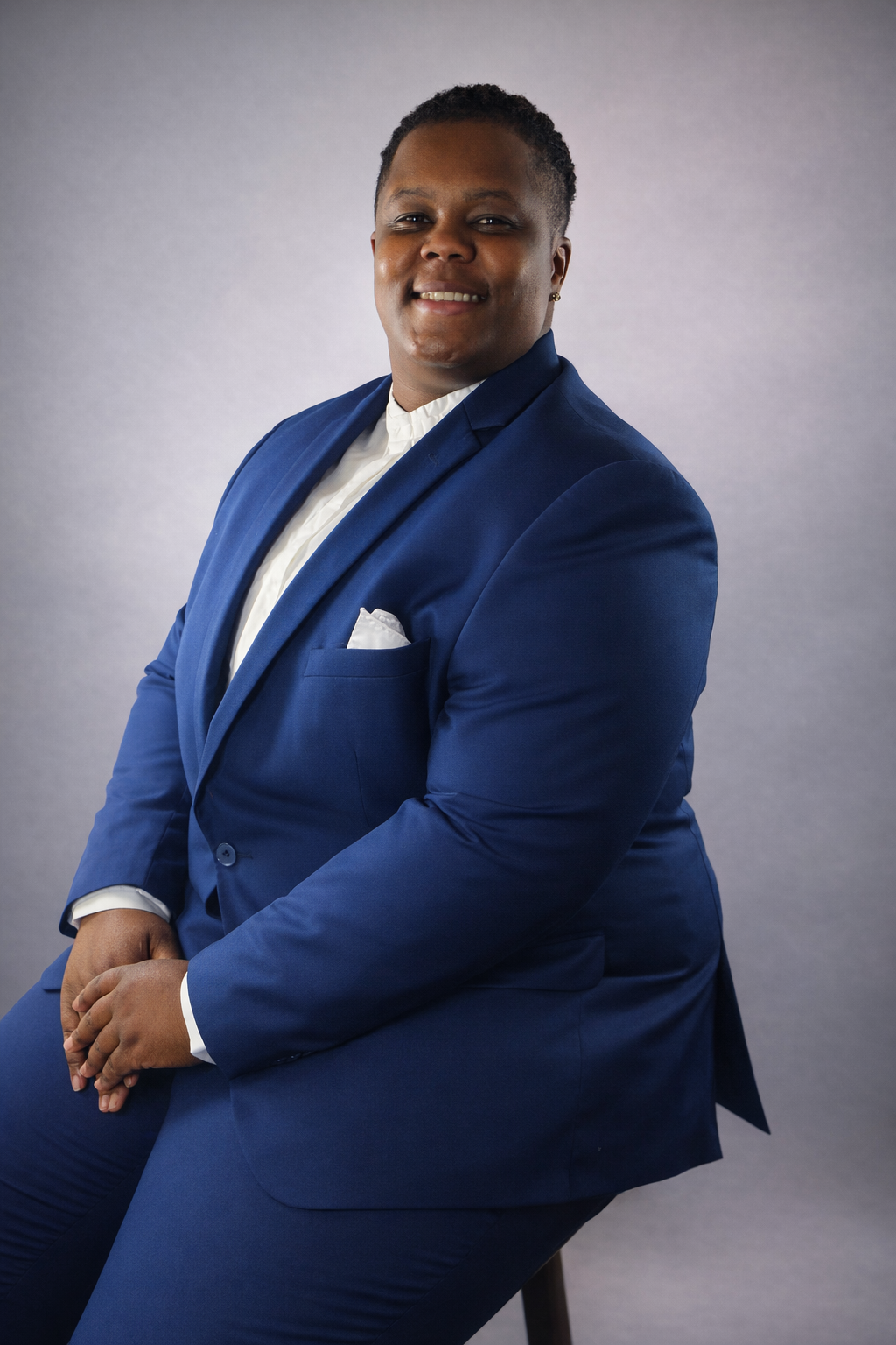 A person in a blue suit and white shirt sitting on a black stool against a gray background, smiling at the camera.