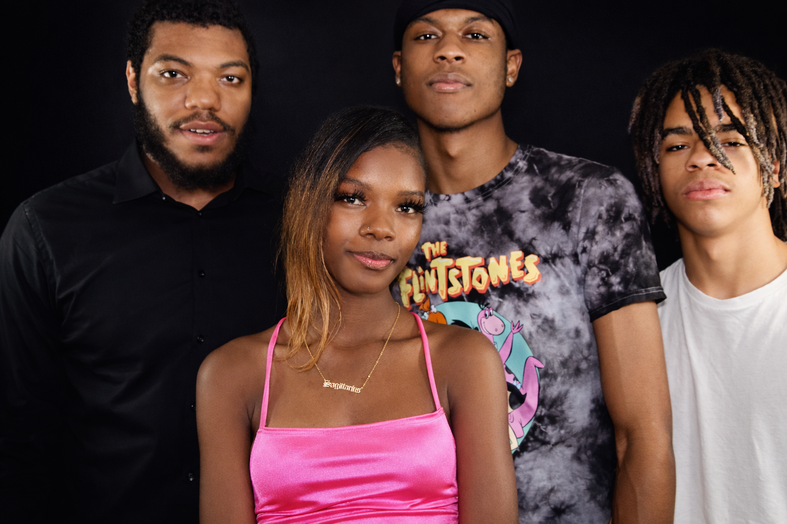 Group of four young adults posing against a black background, with two men on either side and a woman in the center. The woman is wearing a pink satin tank top and a necklace, and the man on the right is wearing a 'The Flintstones' T-shirt. The other