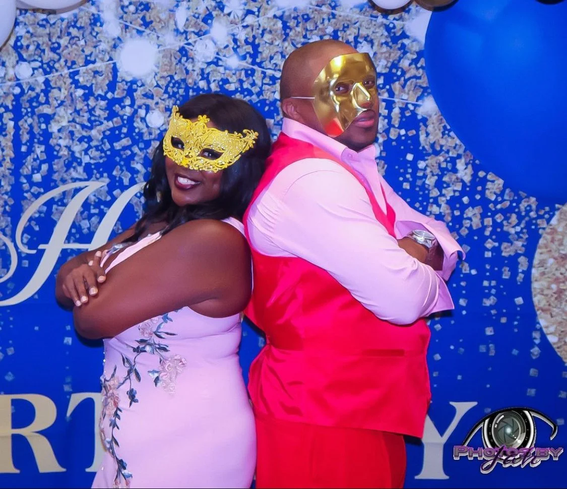 A man and woman standing back to back, wearing colorful masks at a celebration, with decorations and blue balloons in the background.