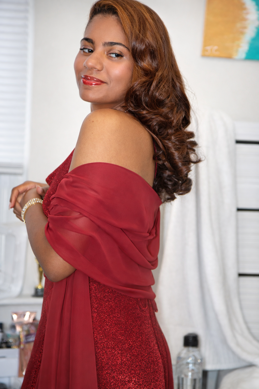 A woman with long, curly brown hair wearing a red dress with a sheer fabric shawl, smiling softly in an indoor setting.