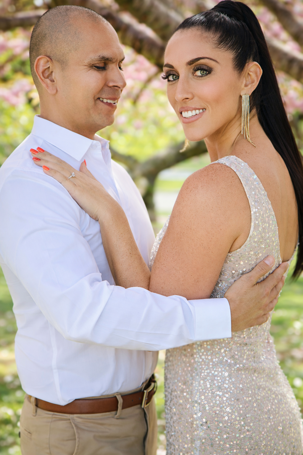 A happy couple stands close together outdoors, smiling at the camera. The woman has long dark hair, bright green eyes, and is wearing a sparkly dress and long earrings. The man has a shaved head, light skin, and is wearing a white shirt with beige pa