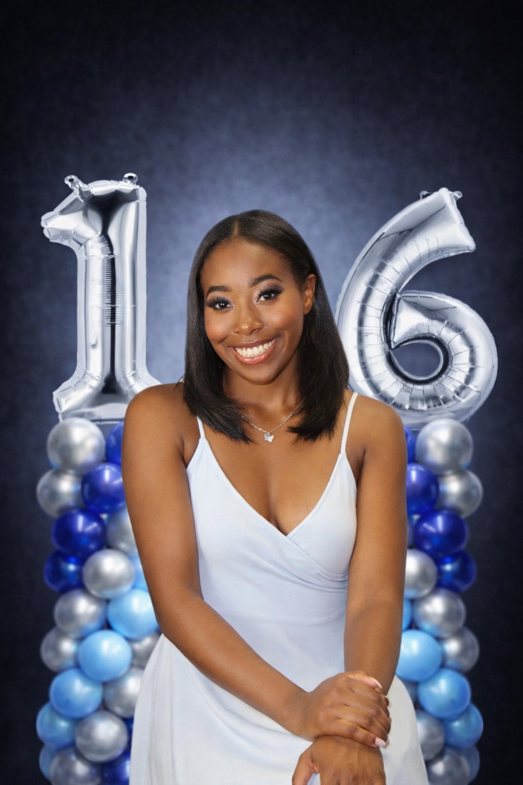 Smiling young woman in a white dress posing in front of balloon numbers 16, with a backdrop of blue and silver balloons.