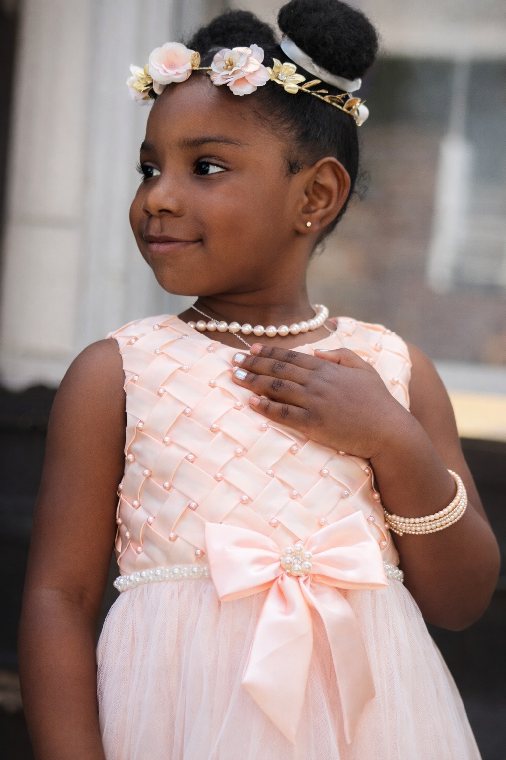 A young girl dressed in a peach-colored dress with pearl embellishments, wearing a pearl necklace, bracelet, and earrings. She has a floral headband with pink and white flowers, and her hair styled in two buns. She has a gentle expression, with her h