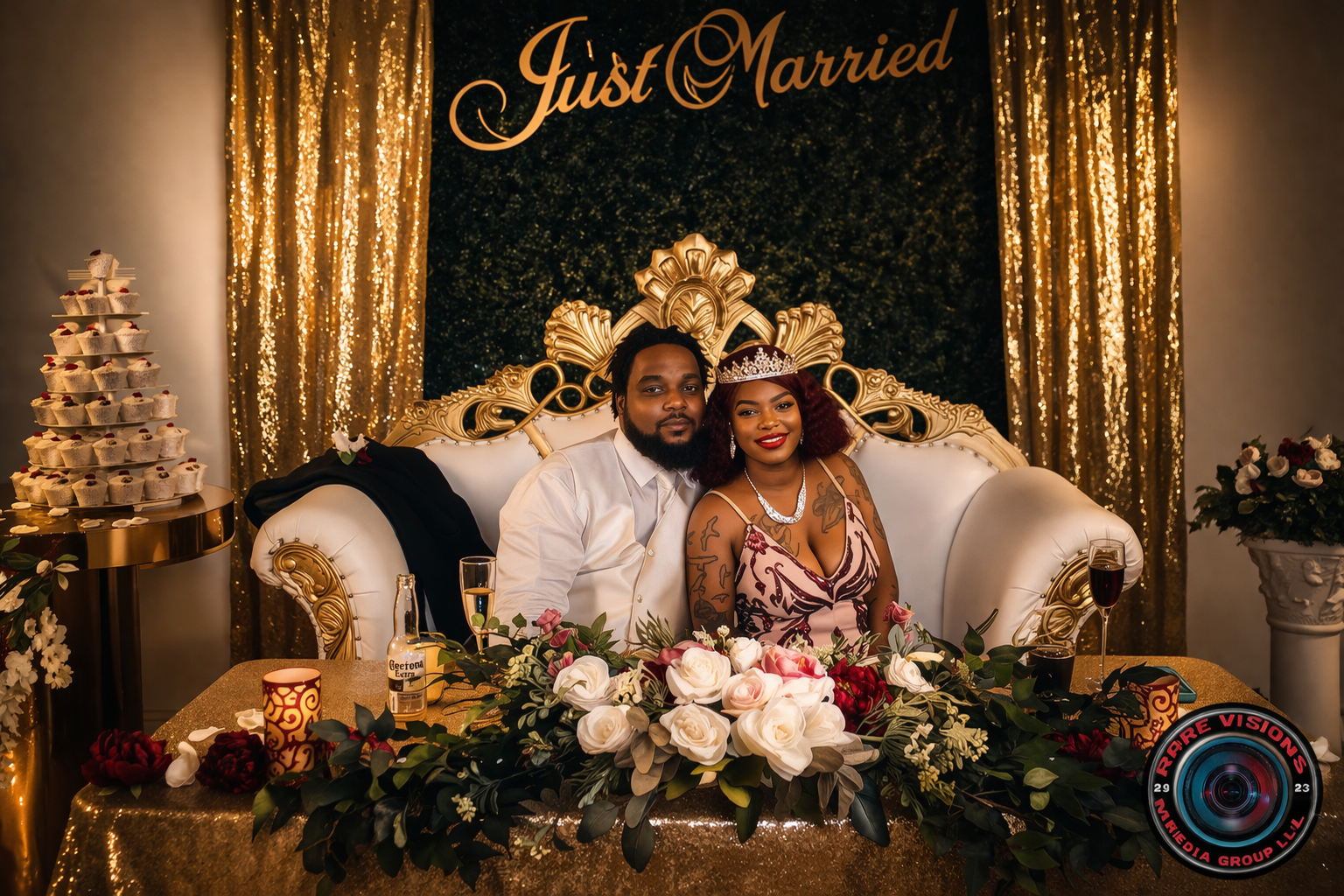 Couple sitting on a decorated couch, celebrating their marriage with a gold and black backdrop that says 'Just Married'. The woman wears a tiara, jewelry, and a floral dress, while the man is in a white shirt. There are flowers, champagne glasses, an