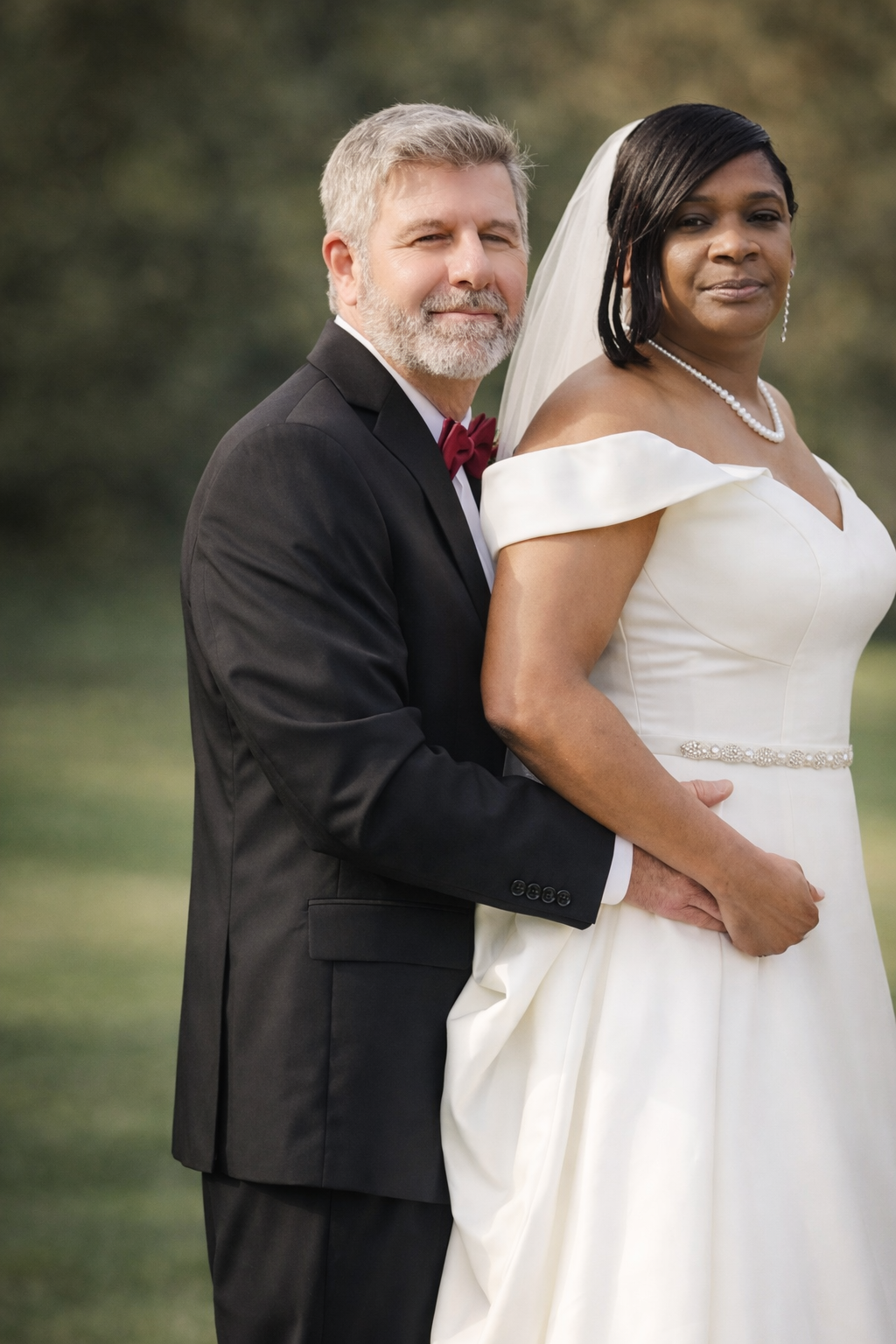A groom with gray hair and beard, dressed in a black tuxedo, stands behind a bride wearing a white wedding gown and veil, outdoors with blurred trees in the background.