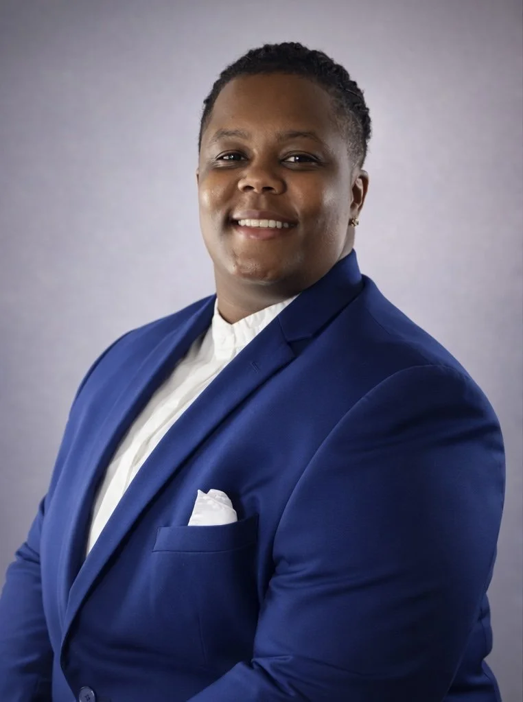 Portrait of a smiling person wearing a blue suit and white shirt against a plain background.