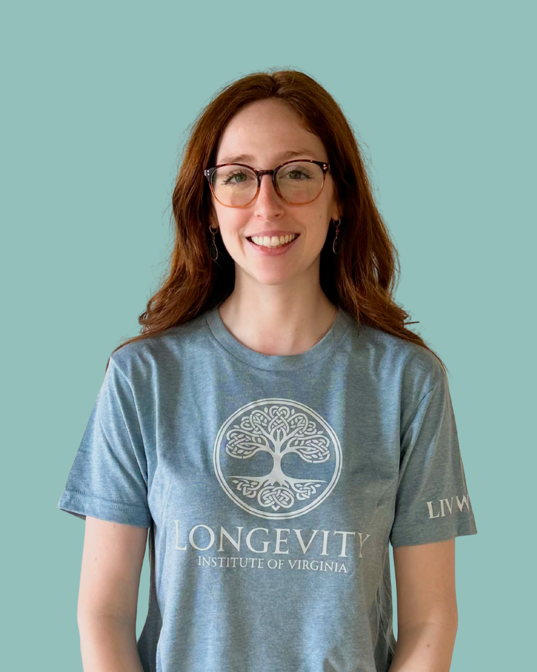 A woman with red hair, glasses, and earrings, smiling, wearing a gray T-shirt with a tree logo and the words 'LONVEVITY INSTITUTE OF VIRGINIA', standing against a teal background.