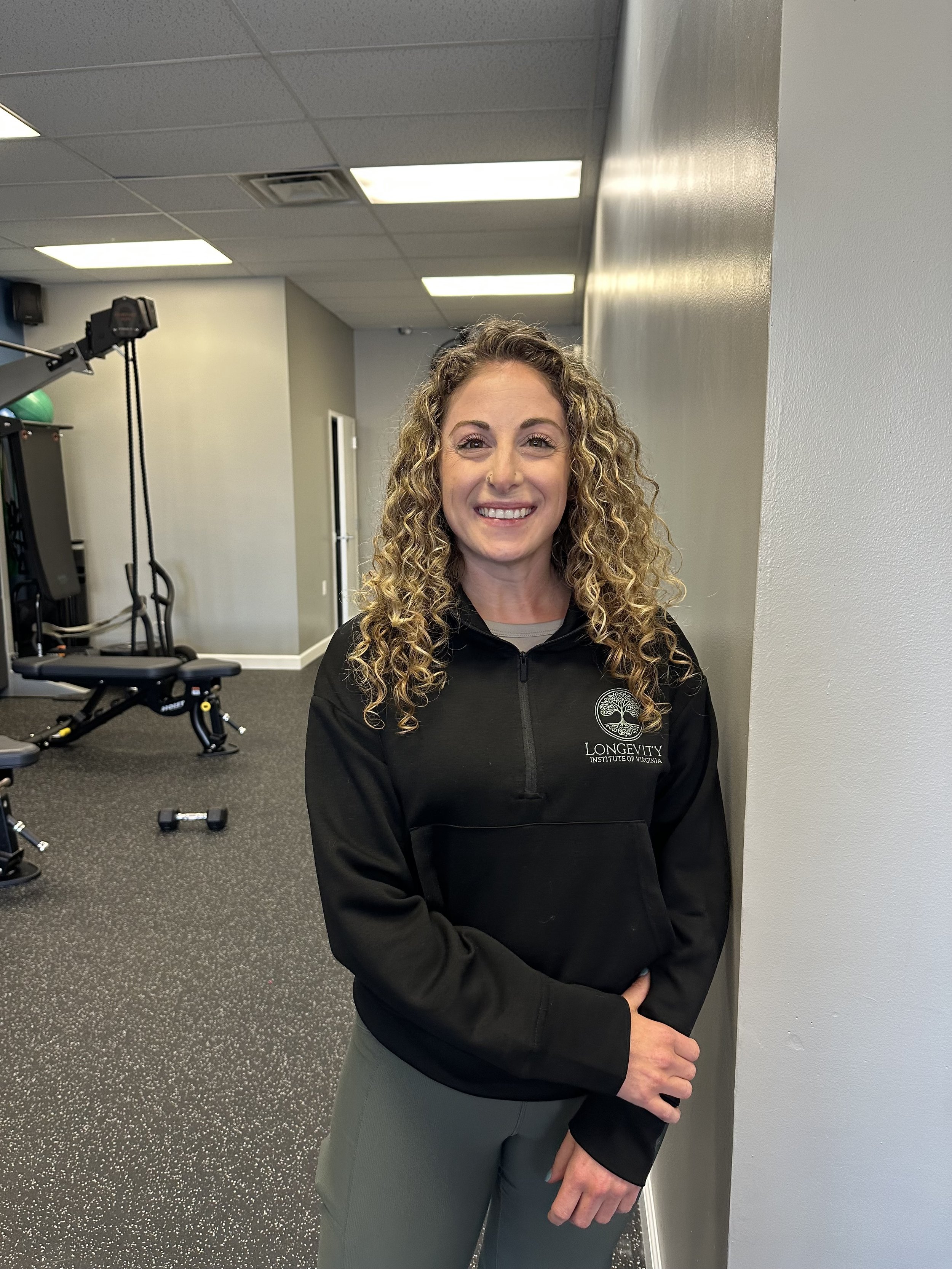 A woman with curly hair smiling, wearing a black jacket with a logo, standing in a room with gym equipment.