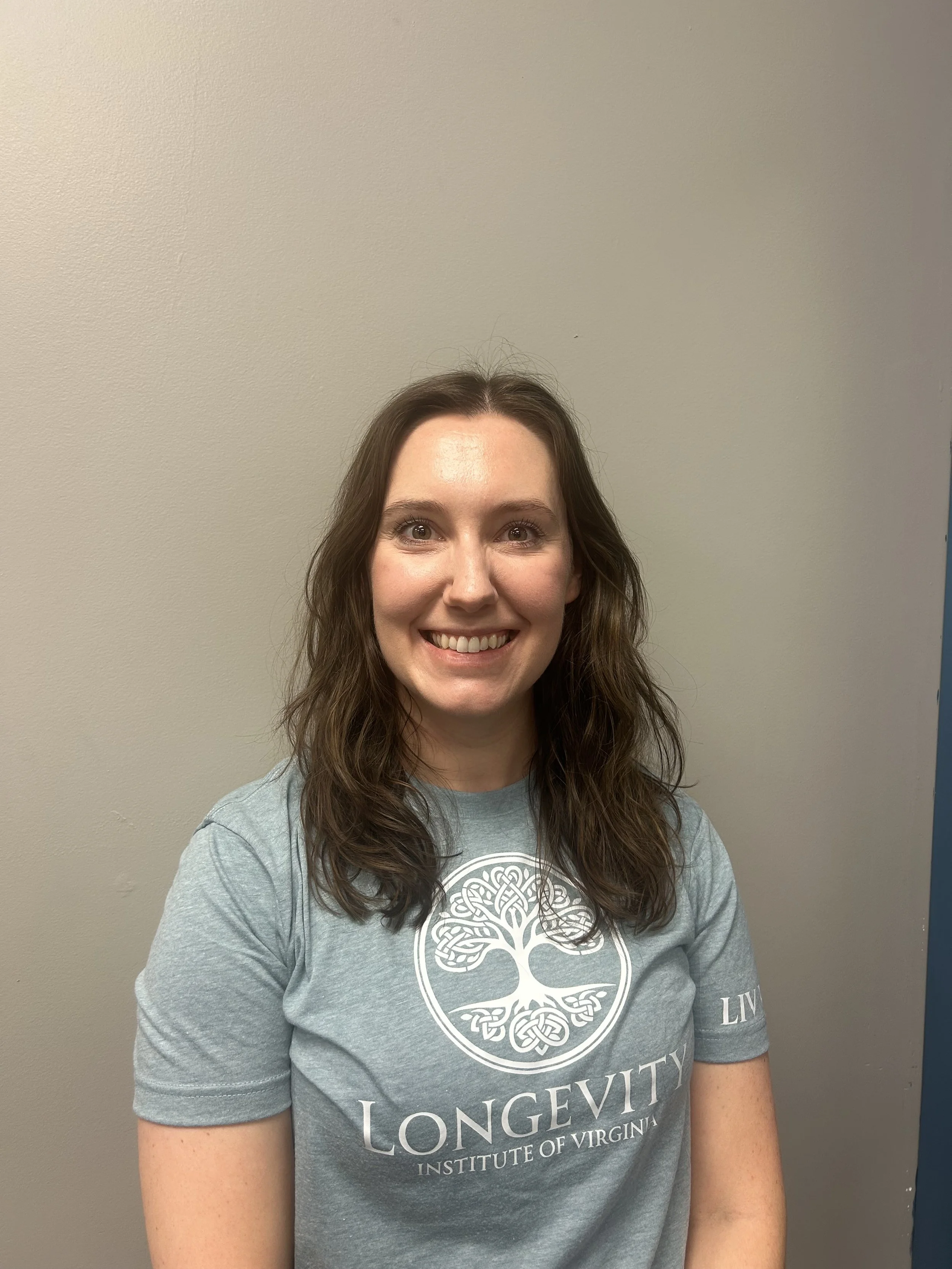 A young woman with brown wavy hair, smiling, wearing a gray t-shirt with a white design and the words 'LONGEVITY INSTITUTE OF VIRGINIA' printed on it, standing against a plain gray wall.