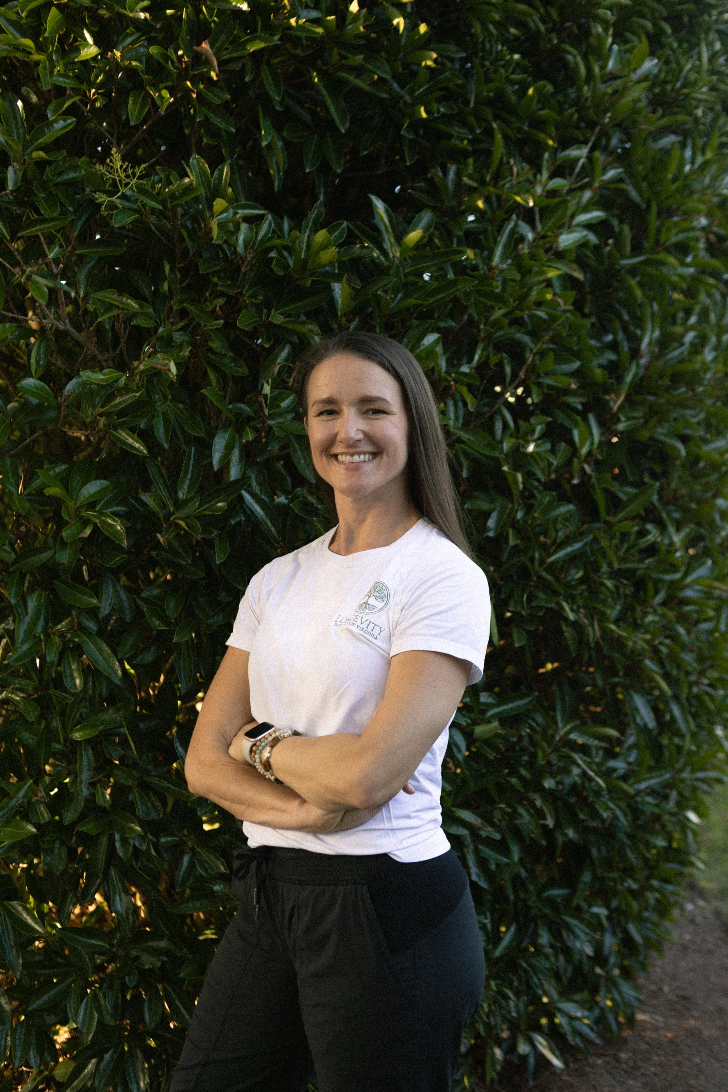 A young woman standing outdoors with arms crossed in front of a large leafy green bush, smiling at the camera, wearing a white T-shirt and black pants.