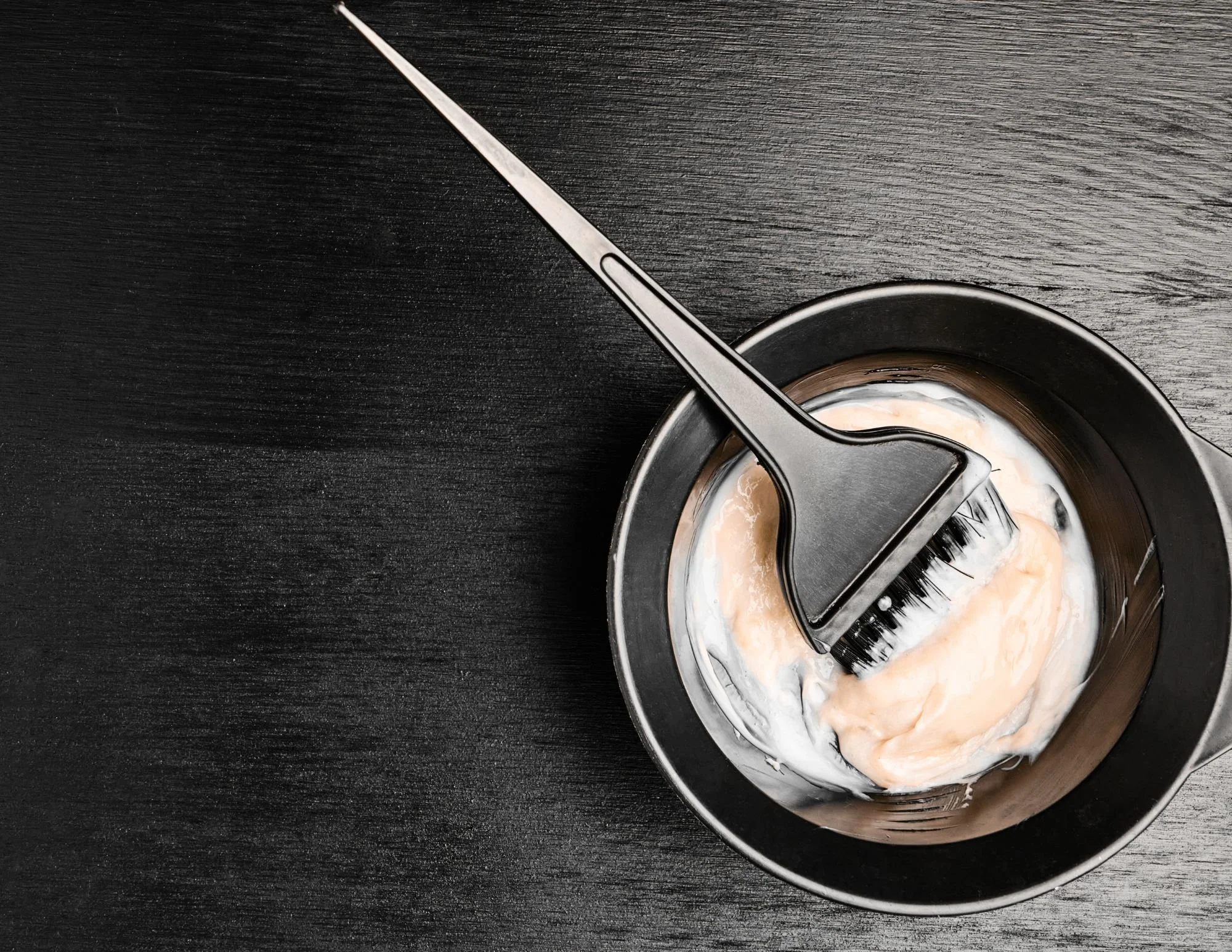 A black mixing bowl with a black brush resting inside, containing a mixture of peach-colored and white substances, placed on a black textured surface.