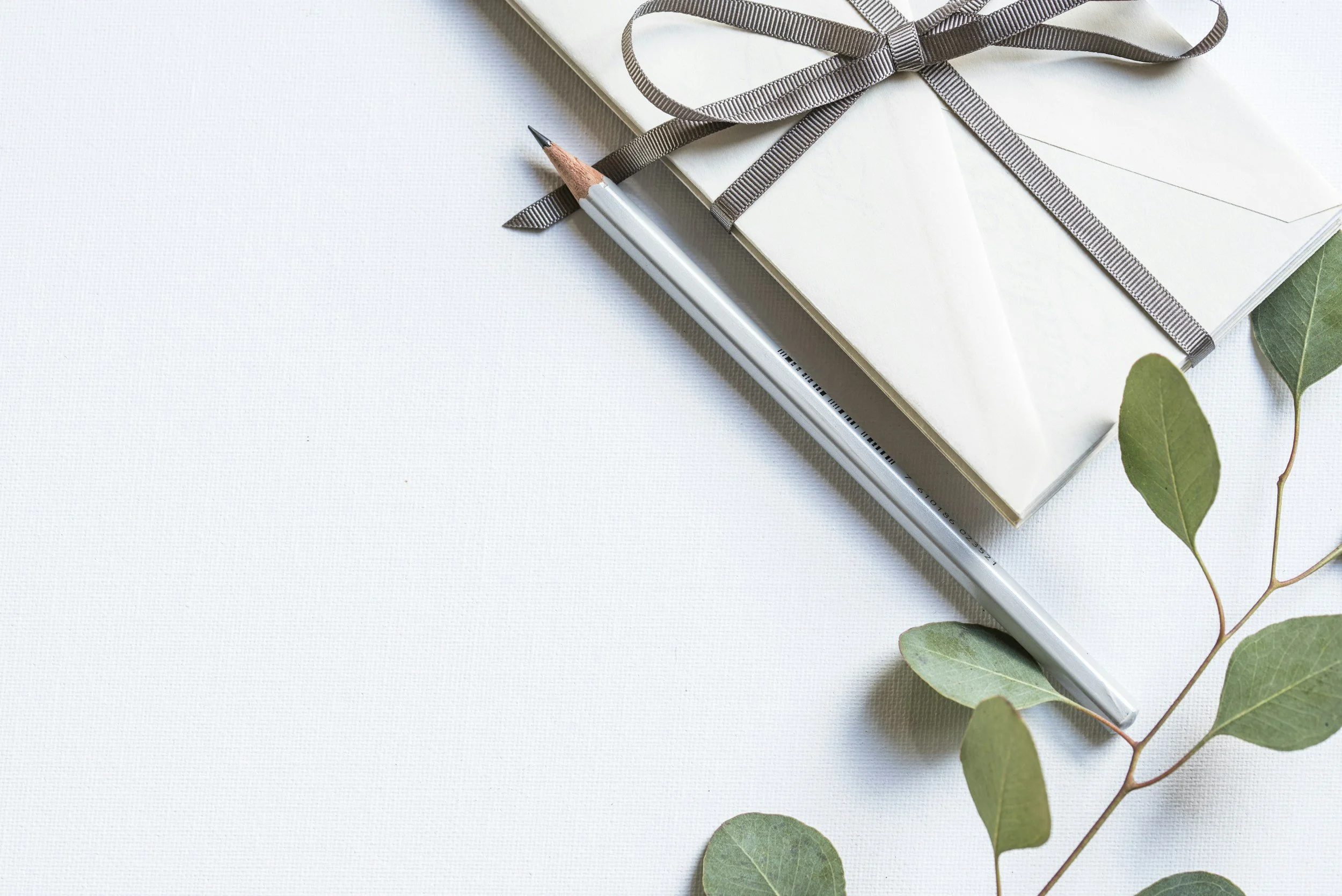 White gift box wrapped with a gray ribbon, a silver pen, and green eucalyptus leaves on a white surface.
