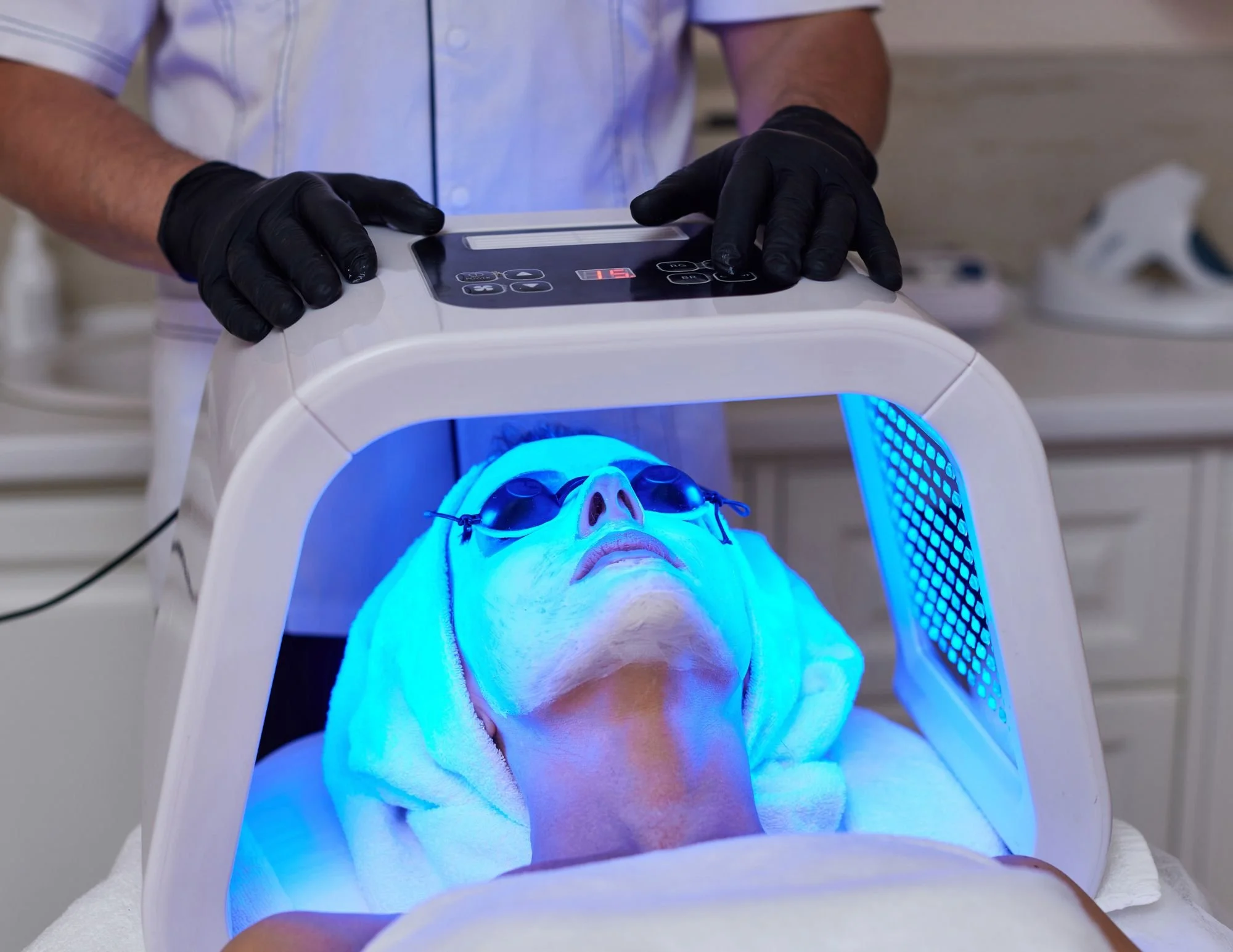 Person lying down with eyes covered by goggles under a blue LED light from a medical device, while a technician in black gloves operates the device.
