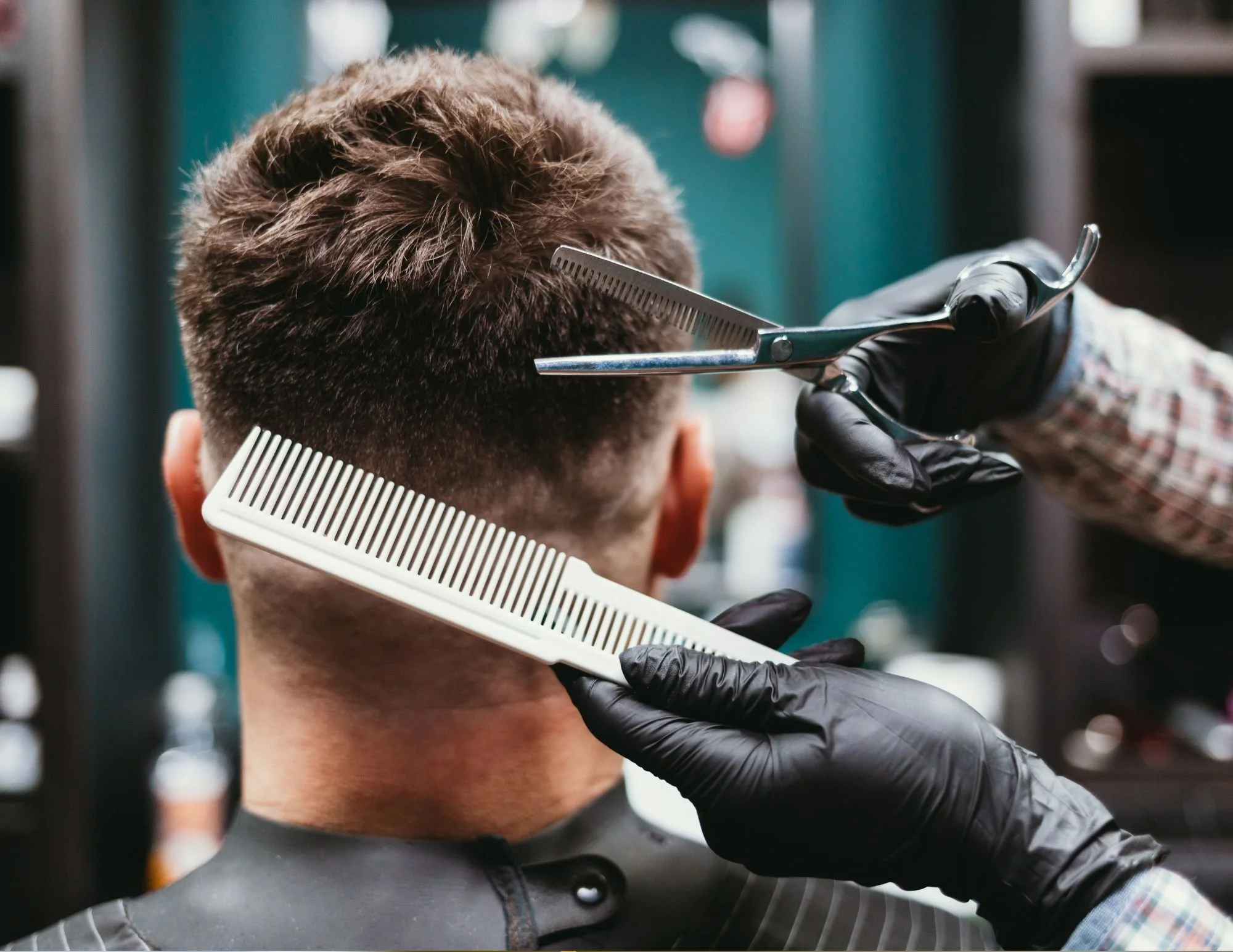A person getting a haircut is sitting in a barber's chair, with a barber using scissors to cut the hair, a comb in the barber's hand, and wearing black gloves.