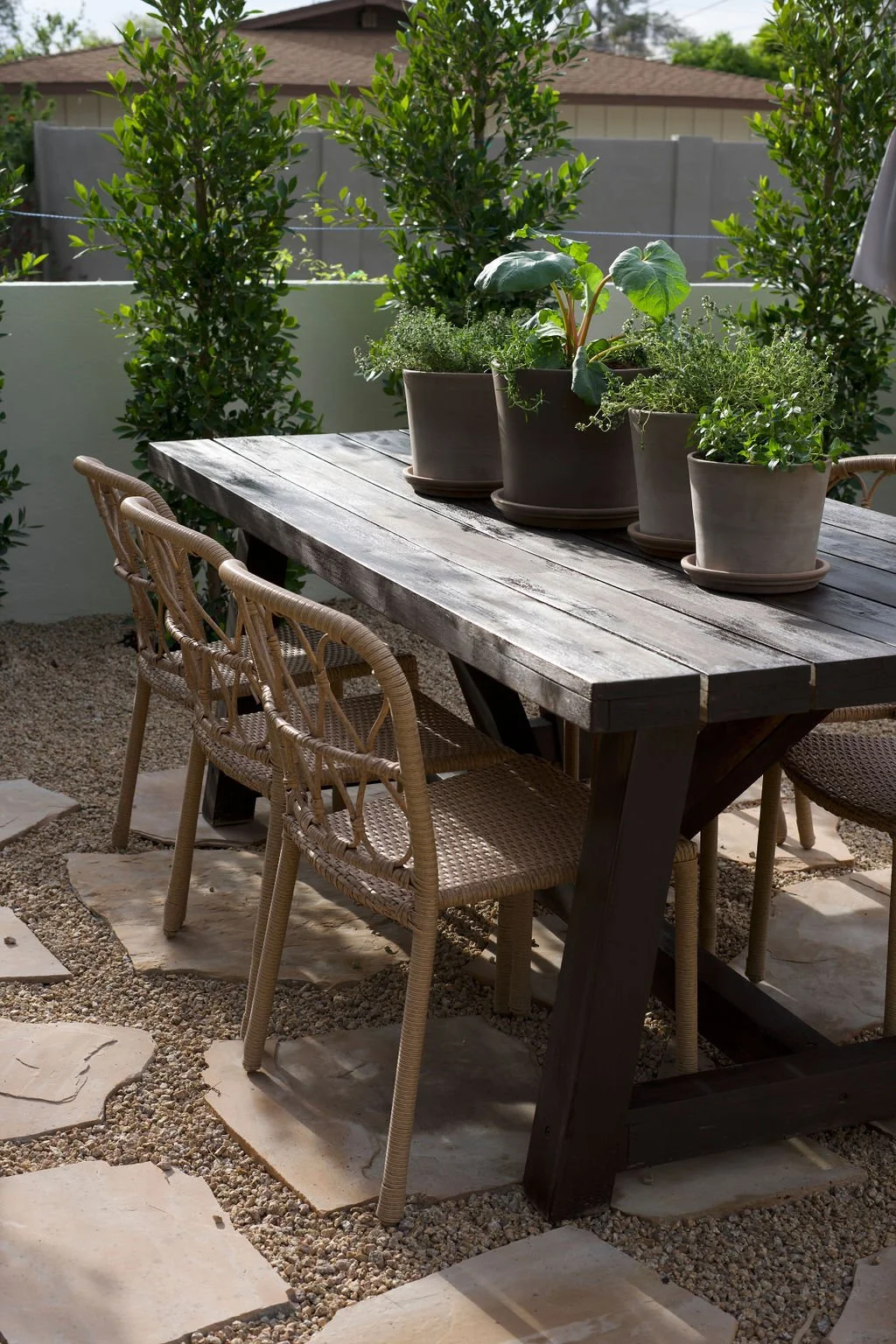 Outdoor patio dining area featuring a rustic wooden table with potted plants on top, surrounded by woven wicker chairs, with a gravel and stone pathway below and lush green shrubs and trees in the background.