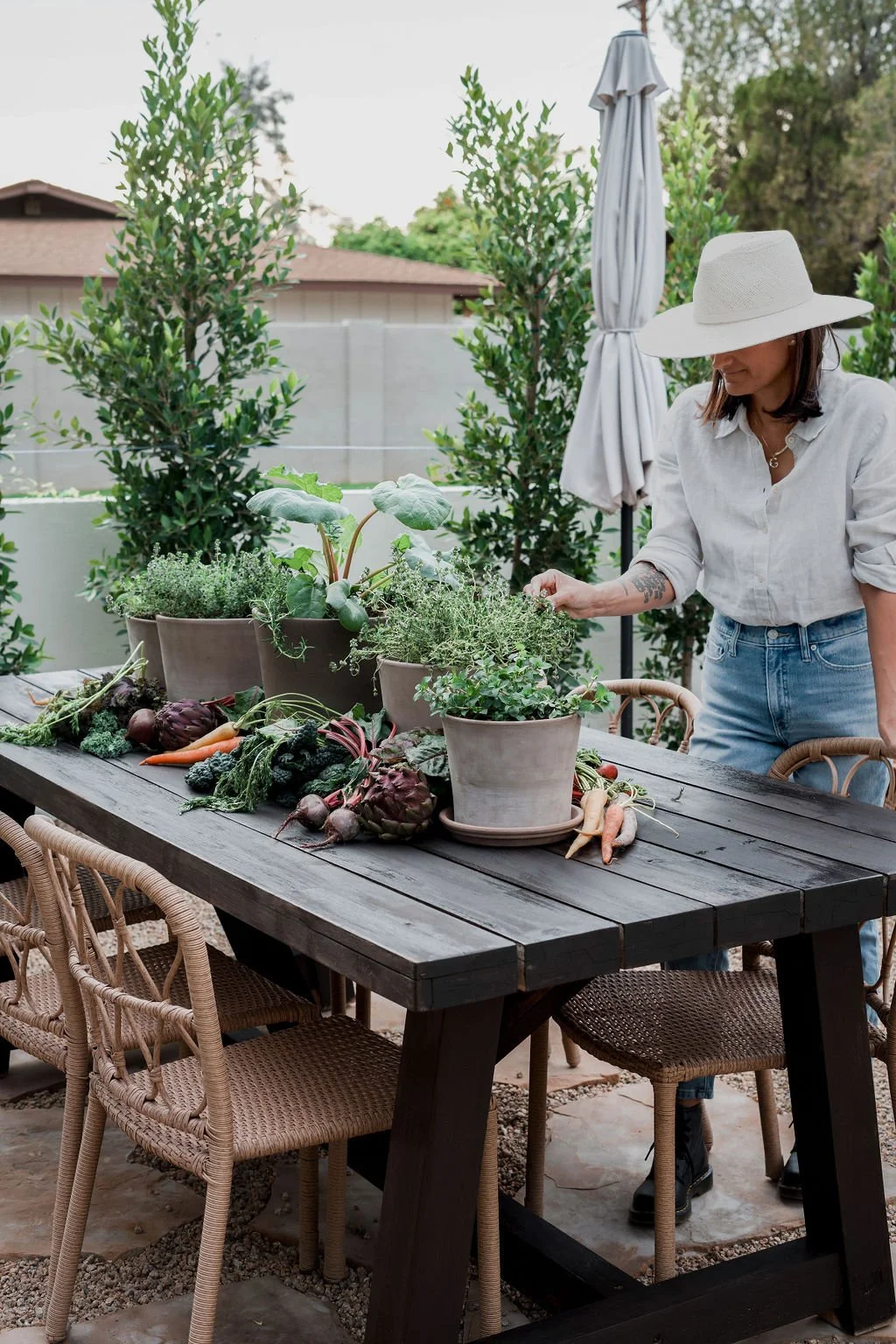 A woman in a white sun hat and white shirt arranging potted herbs and vegetables on a dark wooden outdoor table, with various vegetables and leafy plants, surrounded by green bushes and a backyard.