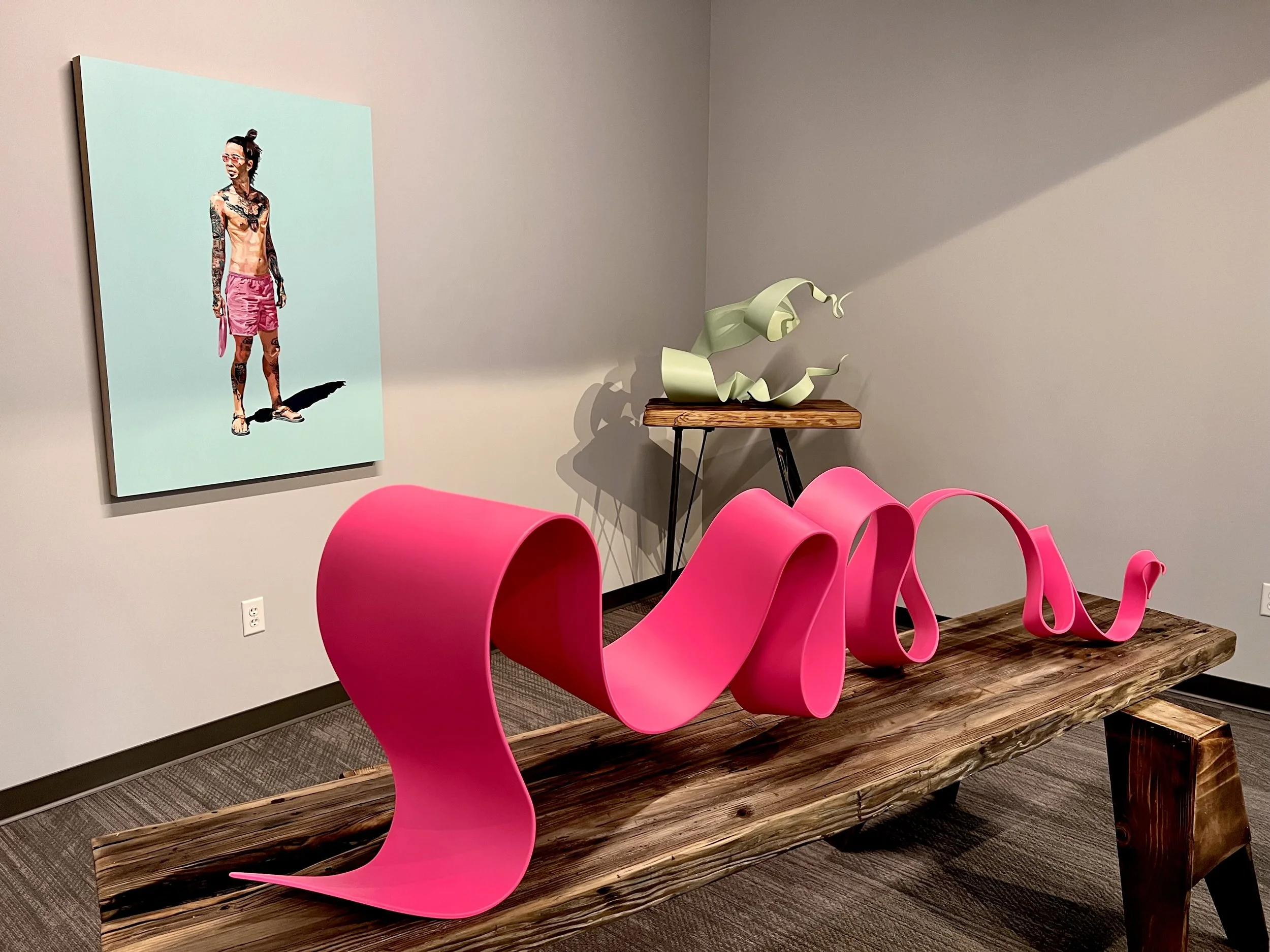 An art gallery with a large pink wavy sculpture on a rustic wooden table, a framed colorful portrait of a tattooed woman on the wall, and a small green abstract sculpture on a side table.