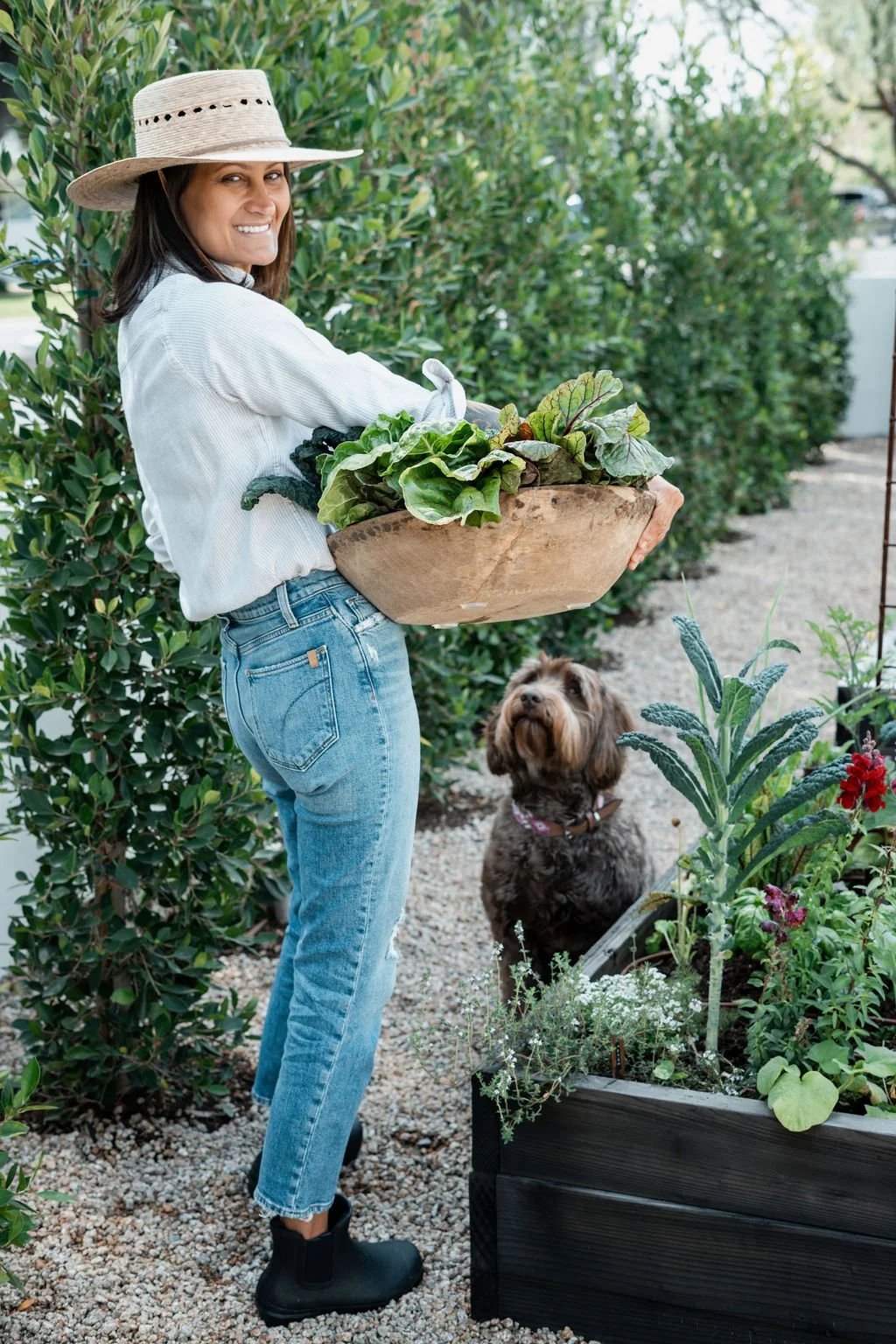 A woman holding a large wooden bowl of leafy greens in a garden, with a brown and tan dog sitting nearby and looking up, surrounded by various plants and flowers.