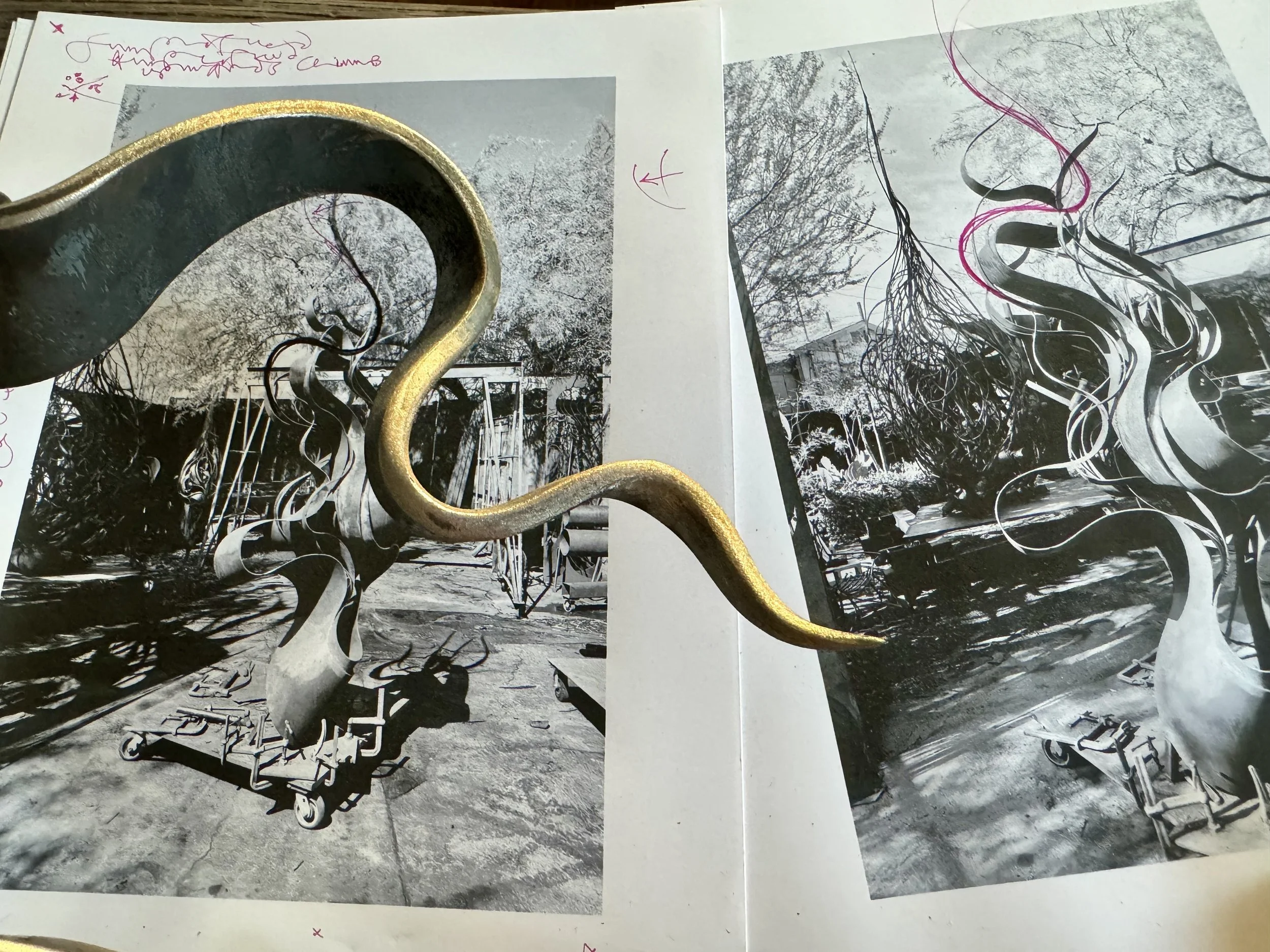 A black and white photo of an outdoor playground area with metal sculptures resembling swirling trees. There is a large, curved metallic sculpture in the foreground partially obscuring the view. The background shows a variety of trees, some structure