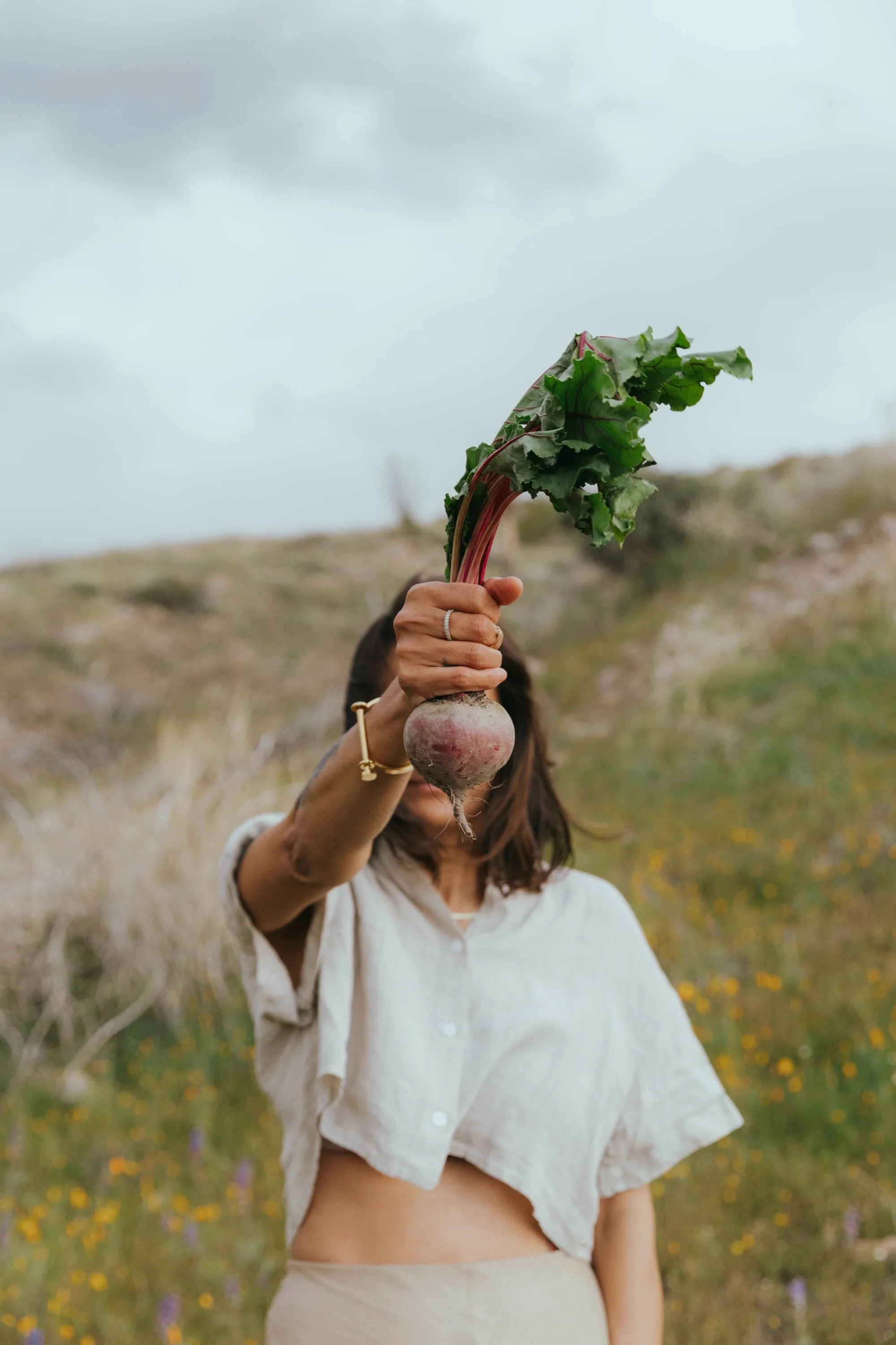 Person holding a large turnip with green leaves, outdoors in a field with wildflowers and a cloudy sky.