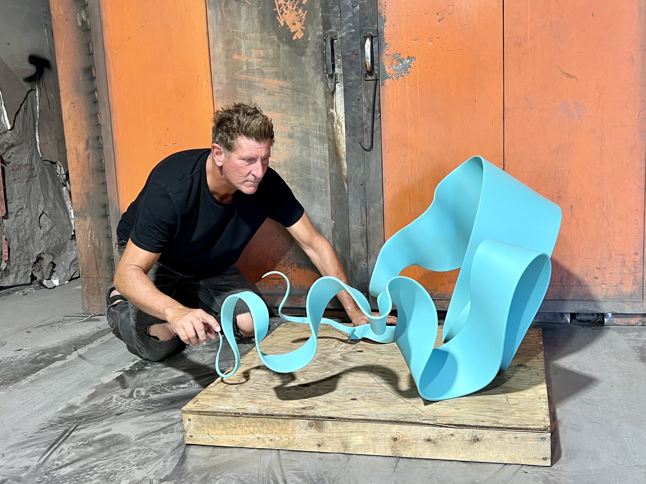 A man wearing a black t-shirt and ripped jeans kneels on the floor next to a large, abstract blue sculpture with flowing curves, set on a wooden platform in an industrial art studio.