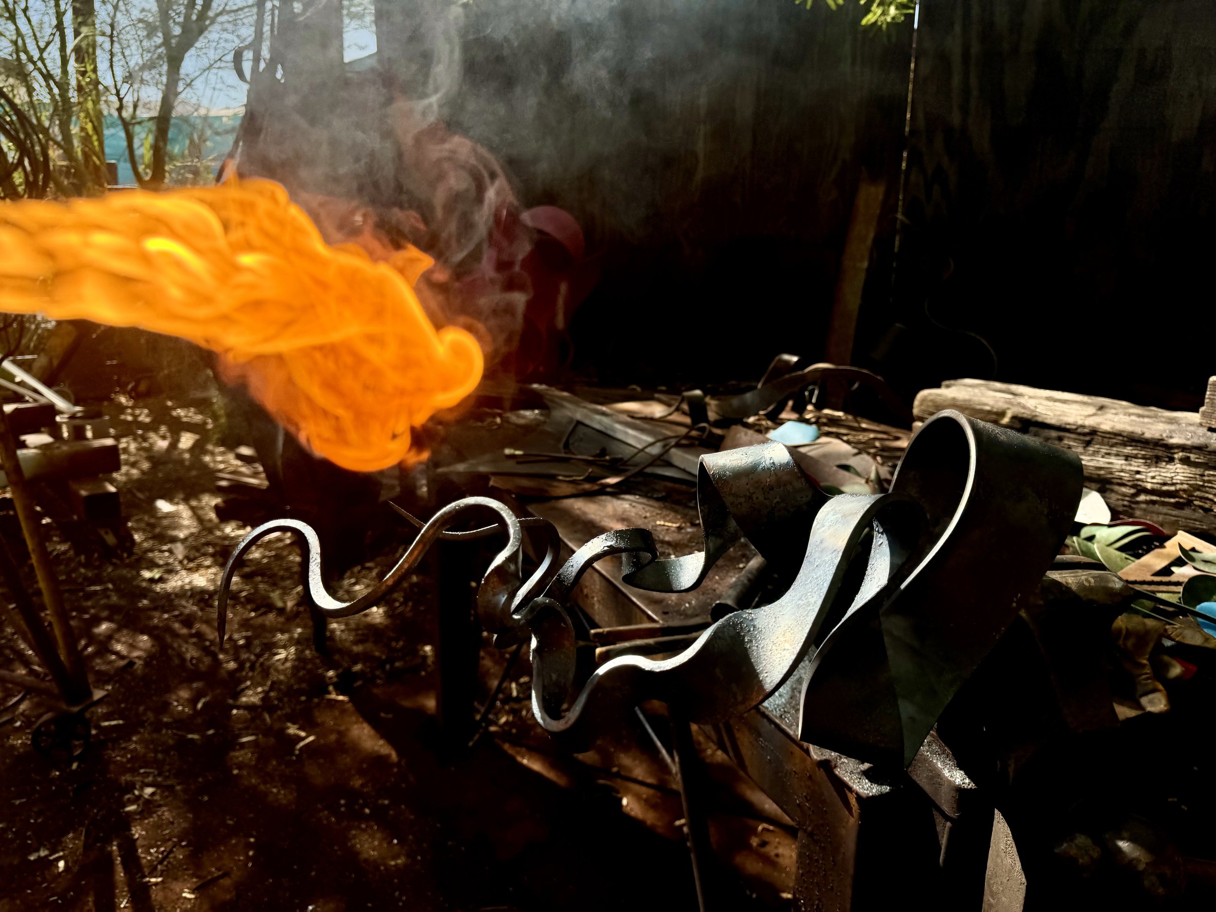 Fire glowing orange with smoke rising, in a blacksmith's forge with metal tools and equipment, with a wooden log and cluttered workspace in the background.