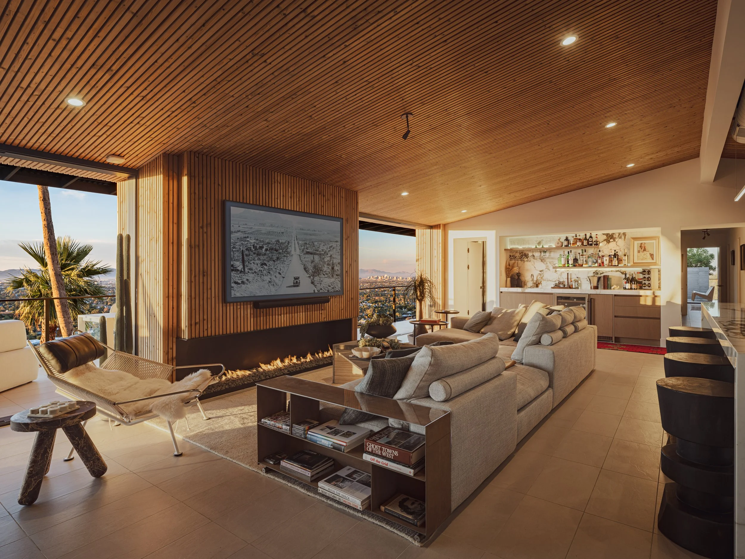 Modern living room with beige sectional sofa, white armchair, wooden ceiling, built-in bar area, large window with city and palm tree view, wall-mounted TV, and a fireplace underneath the TV.