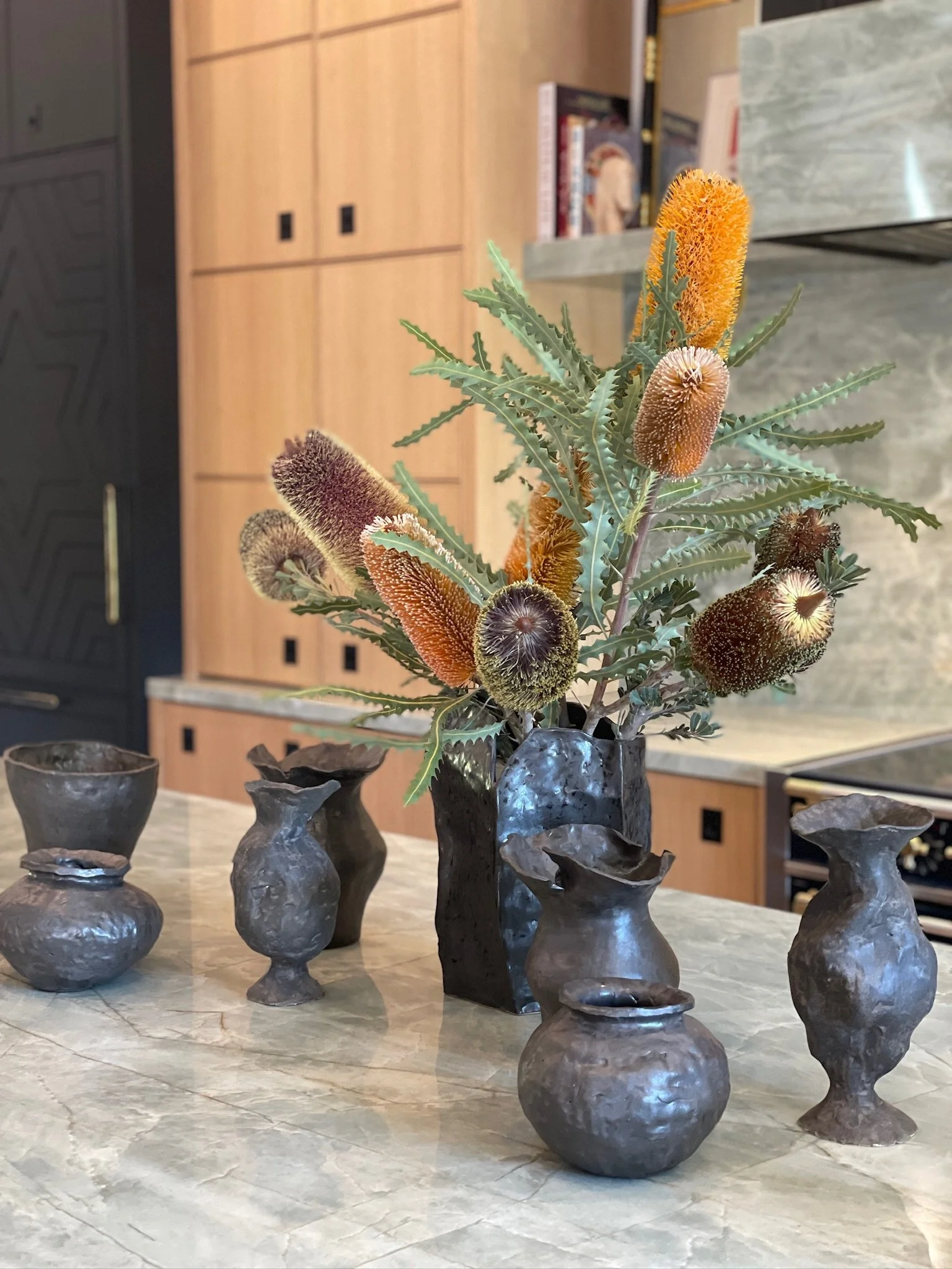 A black vase with a textured surface holds a bouquet of protea flowers and greenery, surrounded by six small, dark, metallic vases on a beige marble table.