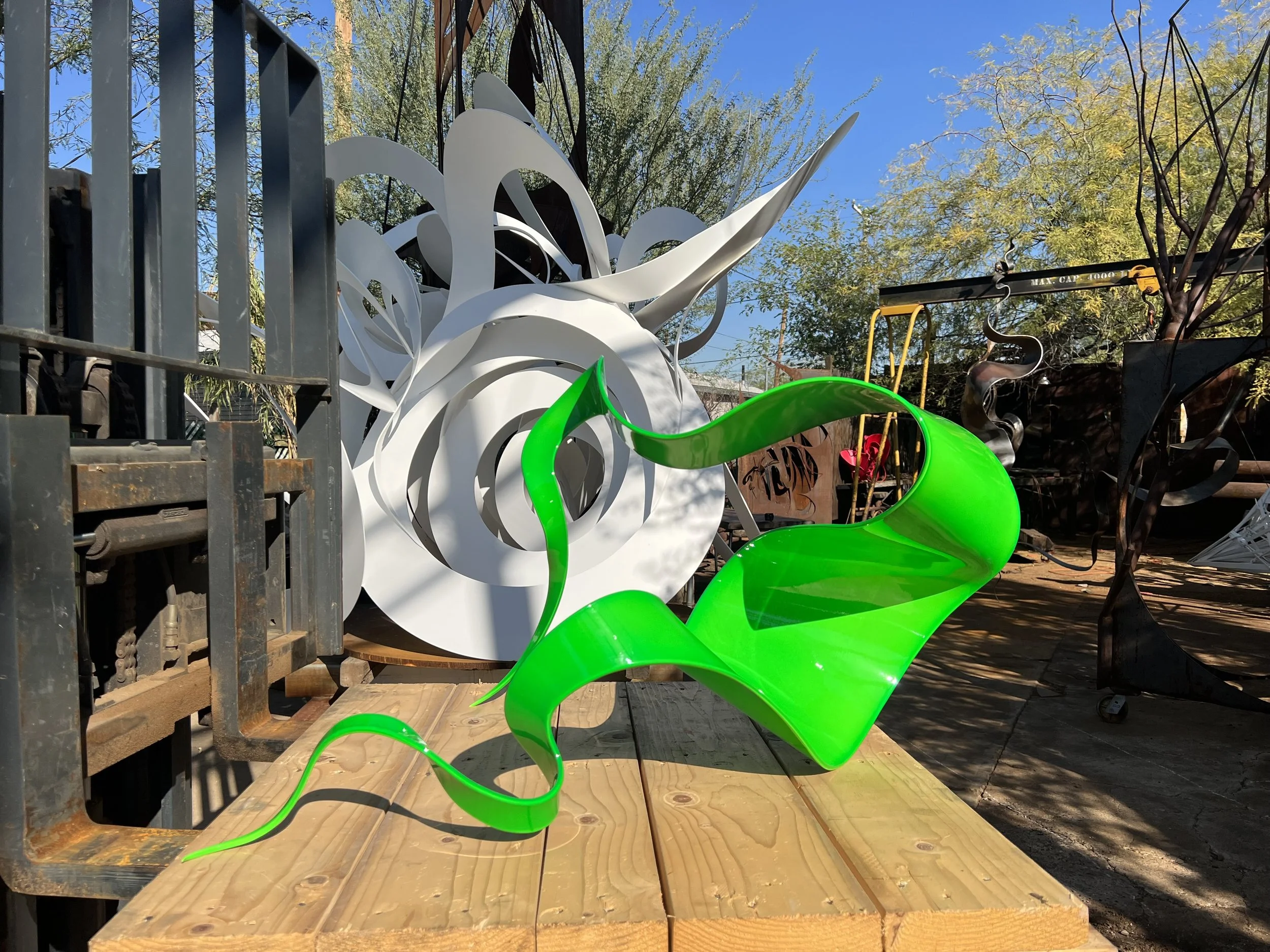 A bright green, wavy, abstract metal sculpture sitting outdoors on a wooden platform, with several large white and black metal sculptures and trees in the background under a clear blue sky.