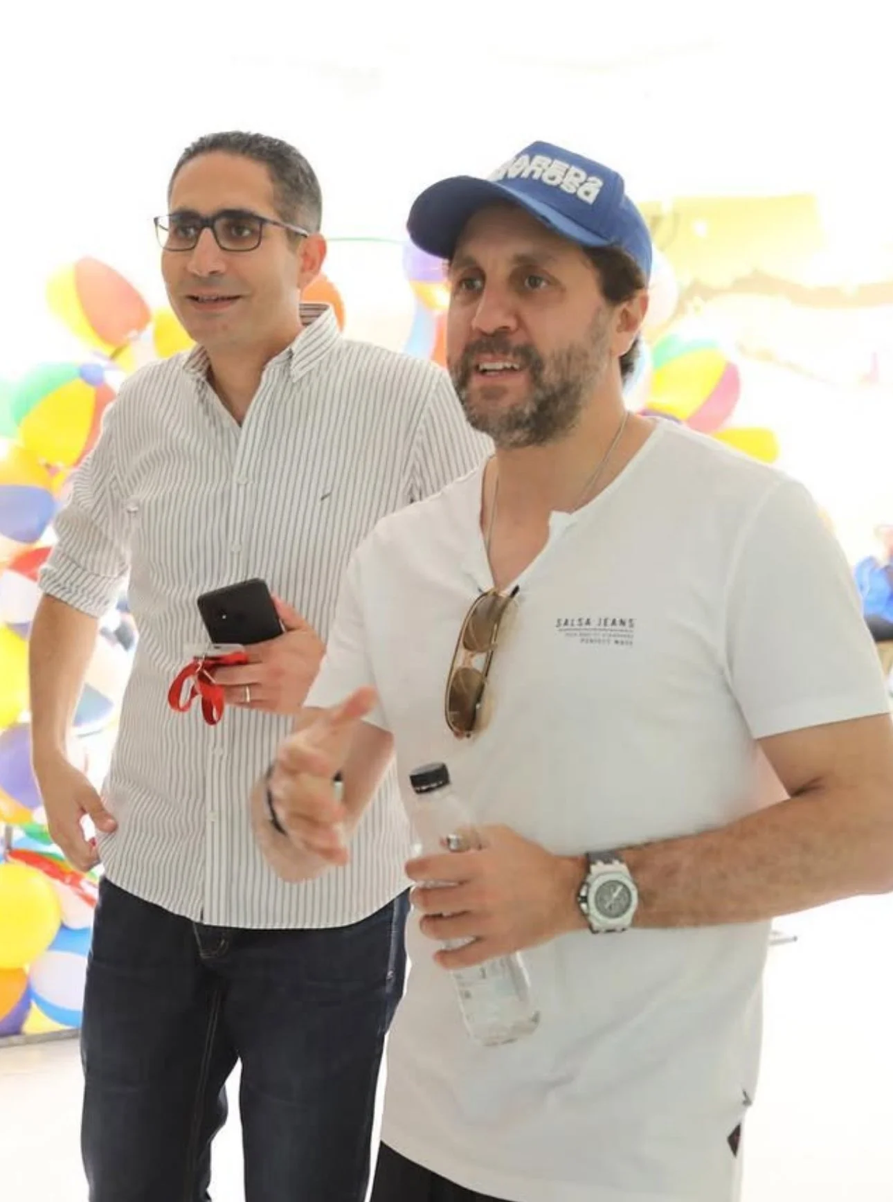 Two men standing together at a colorful event, with balloons in the background. One is wearing glasses and a striped shirt, holding a phone, and the other is wearing a white shirt, sunglasses hanging from his collar, and a blue cap, holding a water bottle.