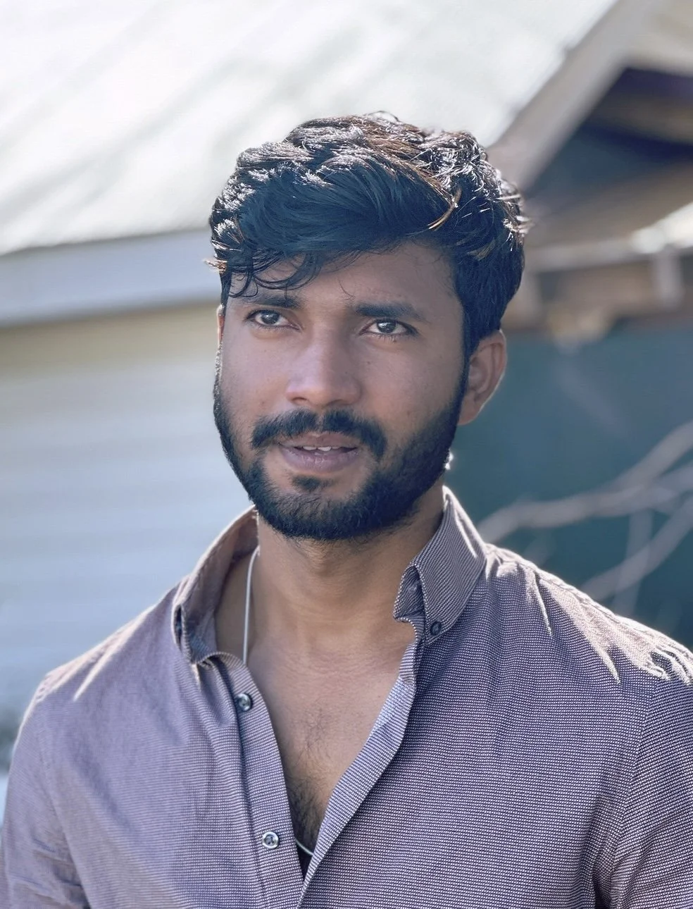 A young man with dark hair and a beard, outdoors, wearing a collared shirt, with a blurred background of a boat and water.