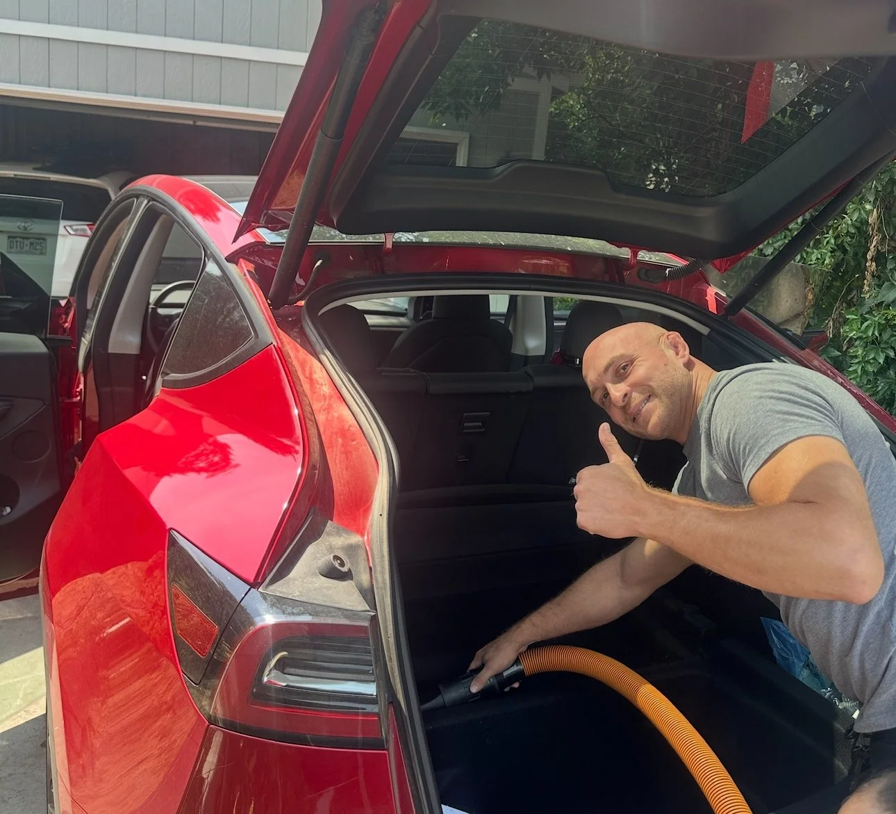 A man smiling and giving a thumbs-up while cleaning the trunk of a red electric vehicle with a vacuum hose.
