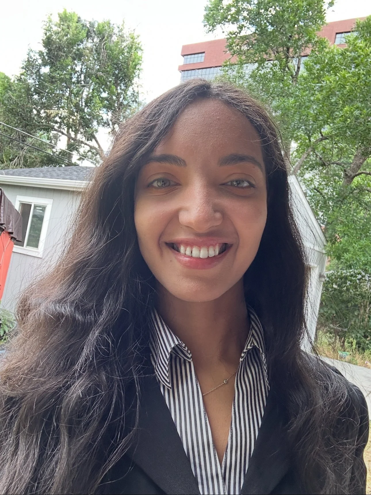 A woman smiling outdoors, wearing a black jacket over a black and white striped shirt, with long dark wavy hair, in a backyard with trees and a building in the background.