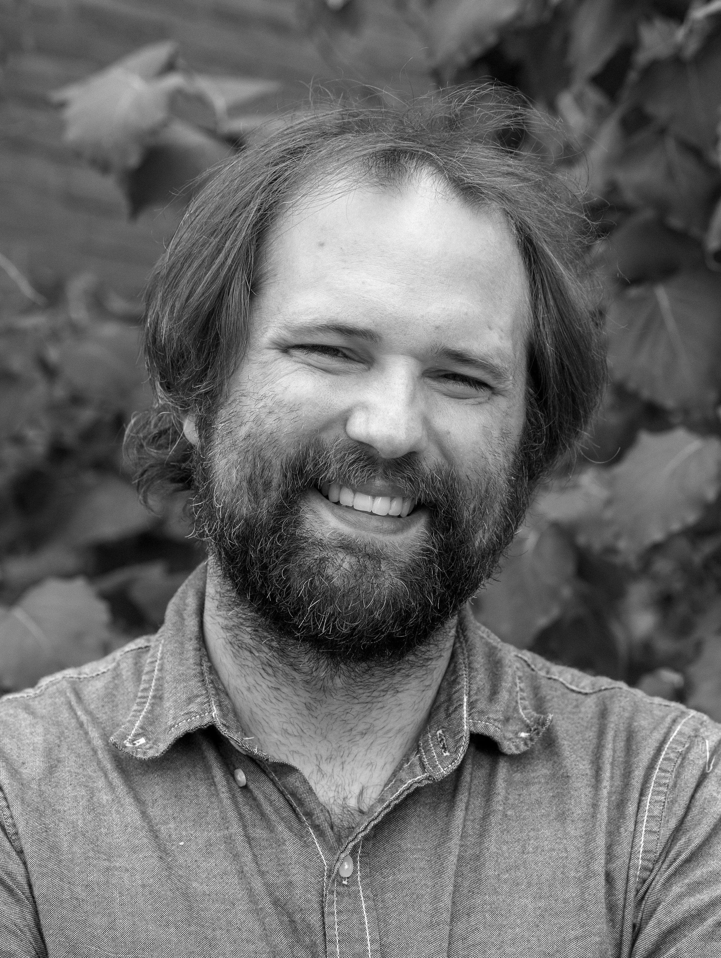 Will Carlson, associate at Ascend Group, with medium-length wavy hair and a short beard, wearing a button-down shirt outdoors.