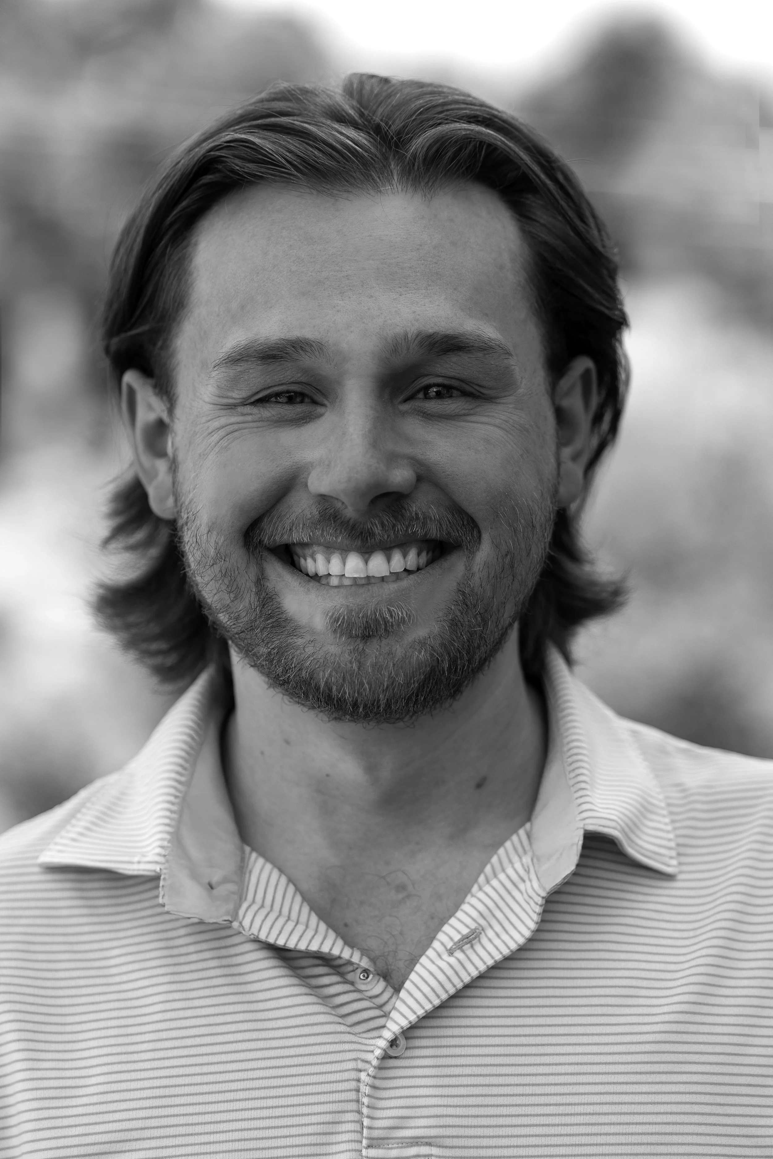Ethan Neer, associate at Ascend Group, with medium-length hair and short beard, wearing a striped collared shirt outdoors.